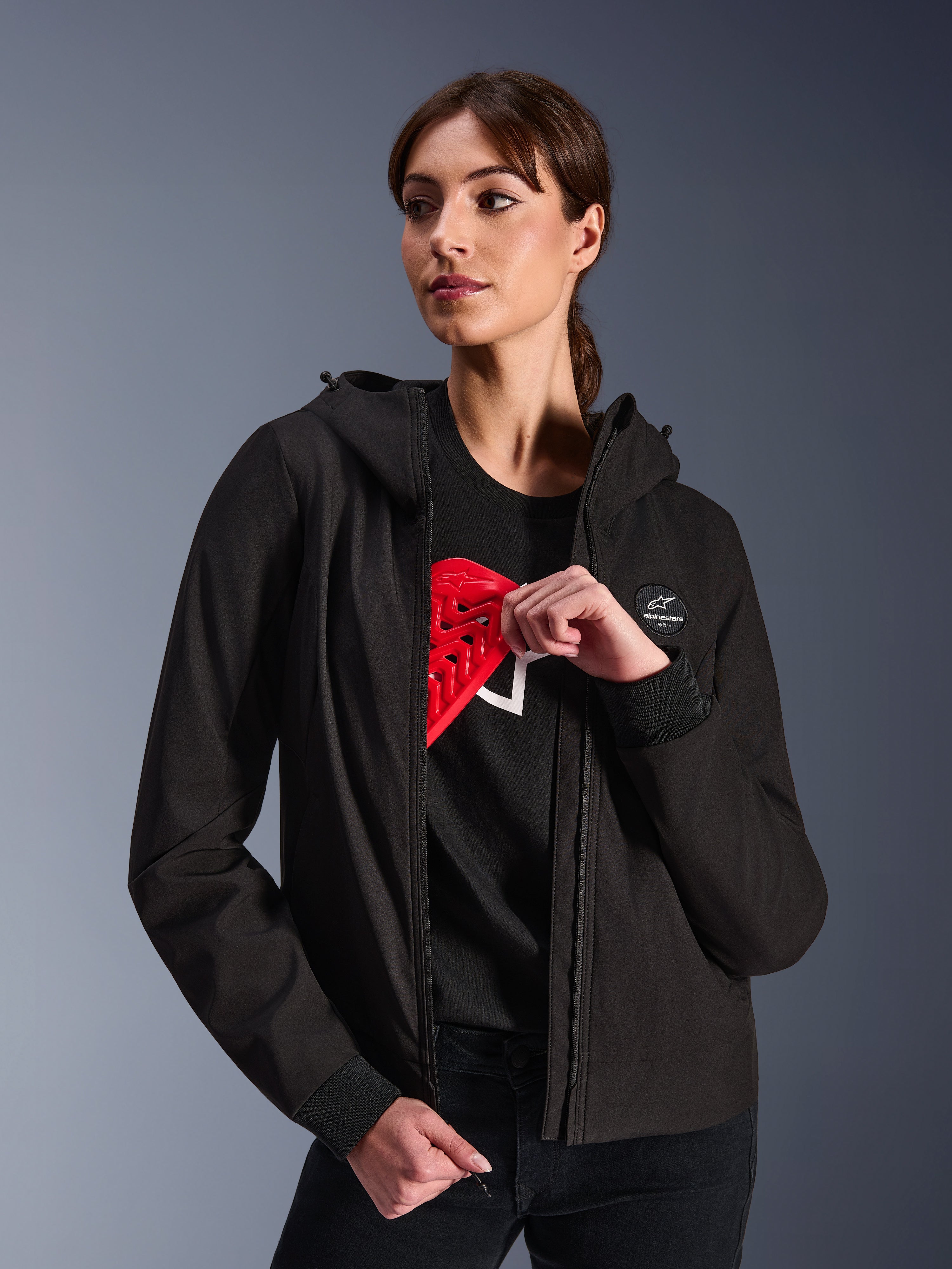 Alpinestars Stella Radium Tech Hoodie voor dames, zwarte urban motorhoodie, vrouwelijk model toont de rode Nucleon Flex Plus-bescherming aan de binnenkant, lichtgewicht 3-laags winddichte textiele buitenlaag met geribde manchetten en logopatch op de borst, beschermende motorkleding