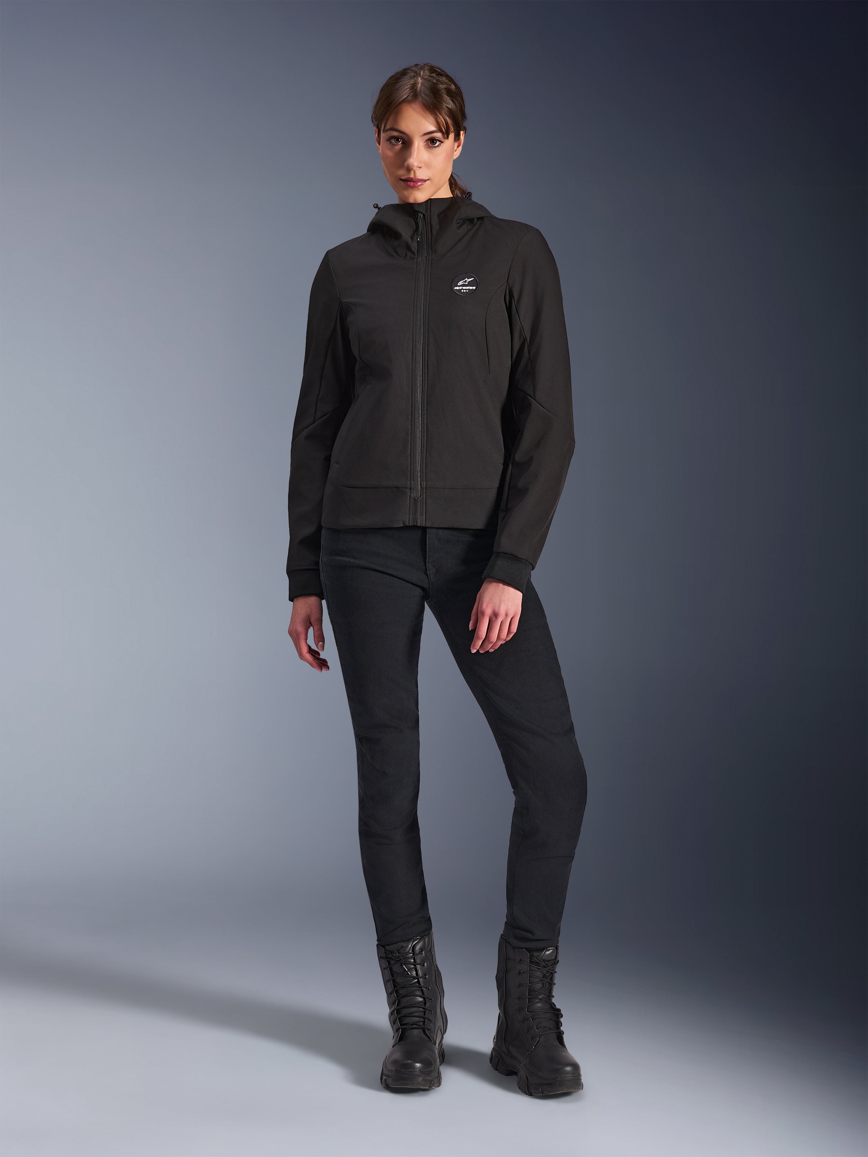 Alpinestars Stella Radium Tech Hoodie in het zwart, urban motor tech shell voor dames, gedragen met een zwarte slim-fit broek en zwarte laarzen, voorzien van een klein logo op de borst en een capuchon, lichtgewicht winddichte constructie voor stedelijk rijden