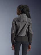 Femme portant le sweat à capuche Alpinestars Stella Radium Tech, vue de dos, Deep Gray Black, coque bicolore grise et noire à 3 couches coupe-vent, logo Alpinestars sur la capuche, vêtement de moto urbain à coupe athlétique