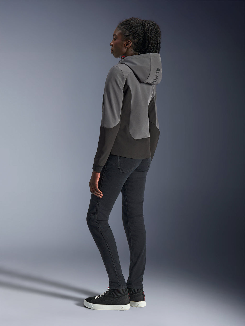 Alpinestars Stella Radium Tech Hoodie pour femme, gris foncé et noir, équipement de moto urbain, vue de dos montrant la capuche avec marquage Alpinestars, porté avec un pantalon slim noir et des baskets montantes
