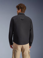 Surchemise Alpinestars Bryon Water Resistant, noir, vue arrière portée par un mannequin, avec logo brodé sur l'empiècement arrière et boucle de suspension utilitaire, associée à un pantalon cargo beige, équipement de moto urbain à l'esthétique workwear classique
