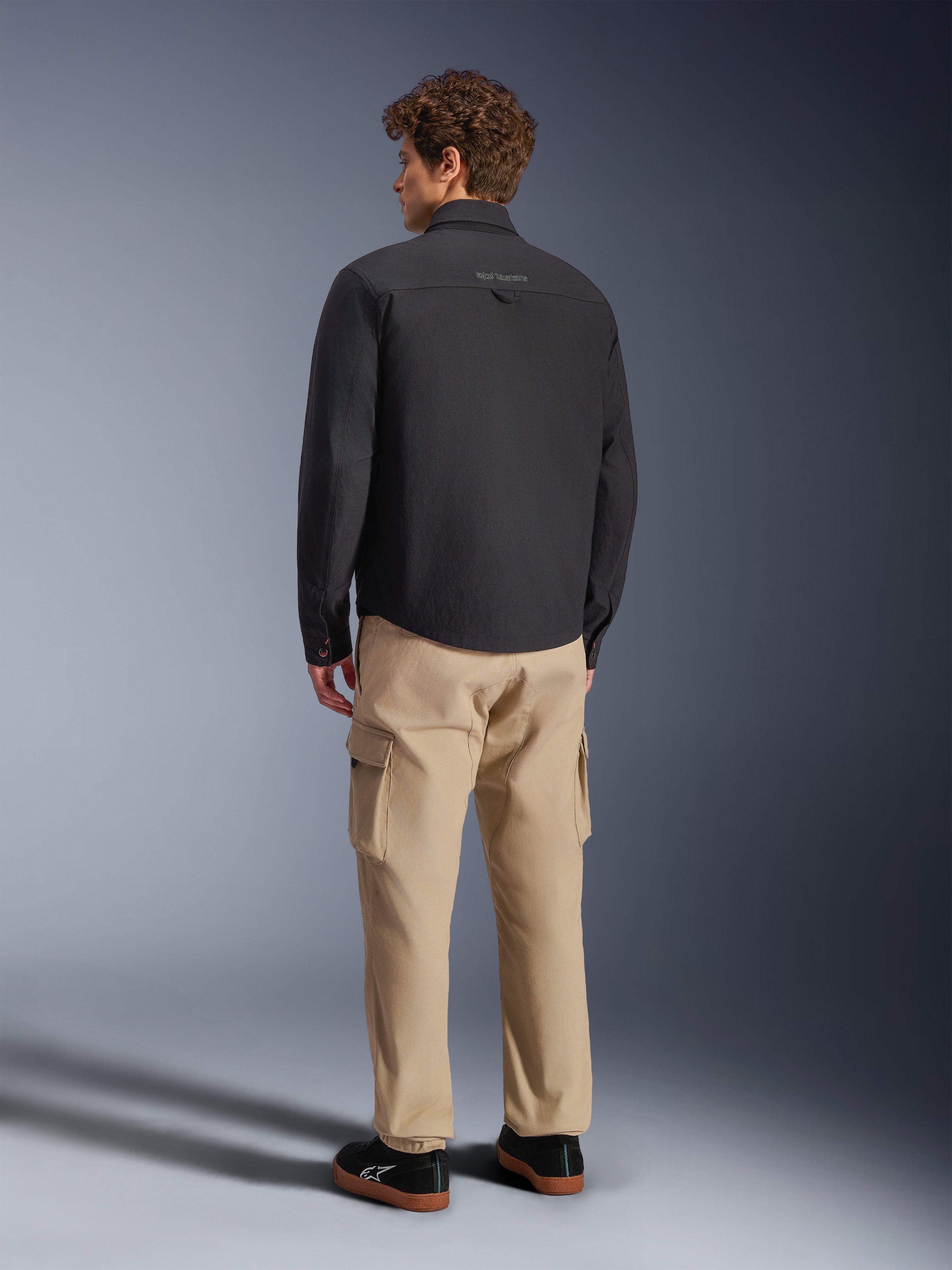 Surchemise Alpinestars Bryon WR, noire, vue de dos sur mannequin, associée à un pantalon cargo beige et des baskets noires, veste de moto urbaine avec texture en toile cirée et design arrière épuré et minimaliste