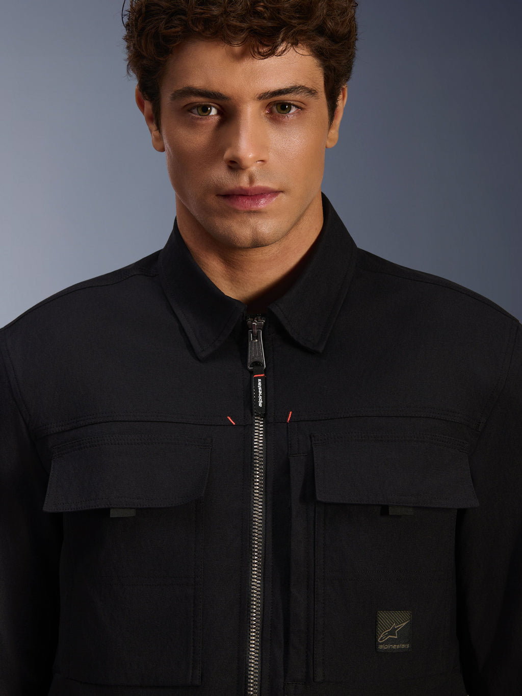 Alpinestars Bryon Water Resistant Overshirt, noire, vue rapprochée de la fermeture éclair frontale et des poches de poitrine, texture en toile cirée durable, design urbain minimaliste avec finitions de la marque et étiquette logo tissée