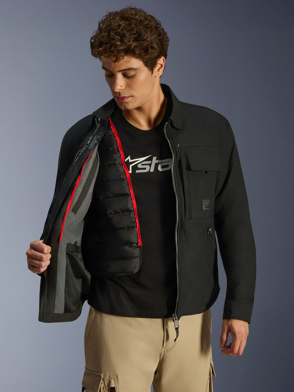 Surchemise Alpinestars Bryon WR, noire, veste de moto urbaine présentée ouverte pour révéler un gilet thermique matelassé noir amovible avec liseré rouge, portée avec un pantalon cargo beige et un t-shirt à logo, design fonctionnel d'inspiration workwear