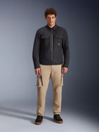 Modèle masculin portant la surchemise résistante à l'eau Alpinestars Bryon noire, dotée de poches cargo sur la poitrine et d'une fermeture éclair centrale, associée à un pantalon cargo beige et des baskets noires, portrait de studio en pied.