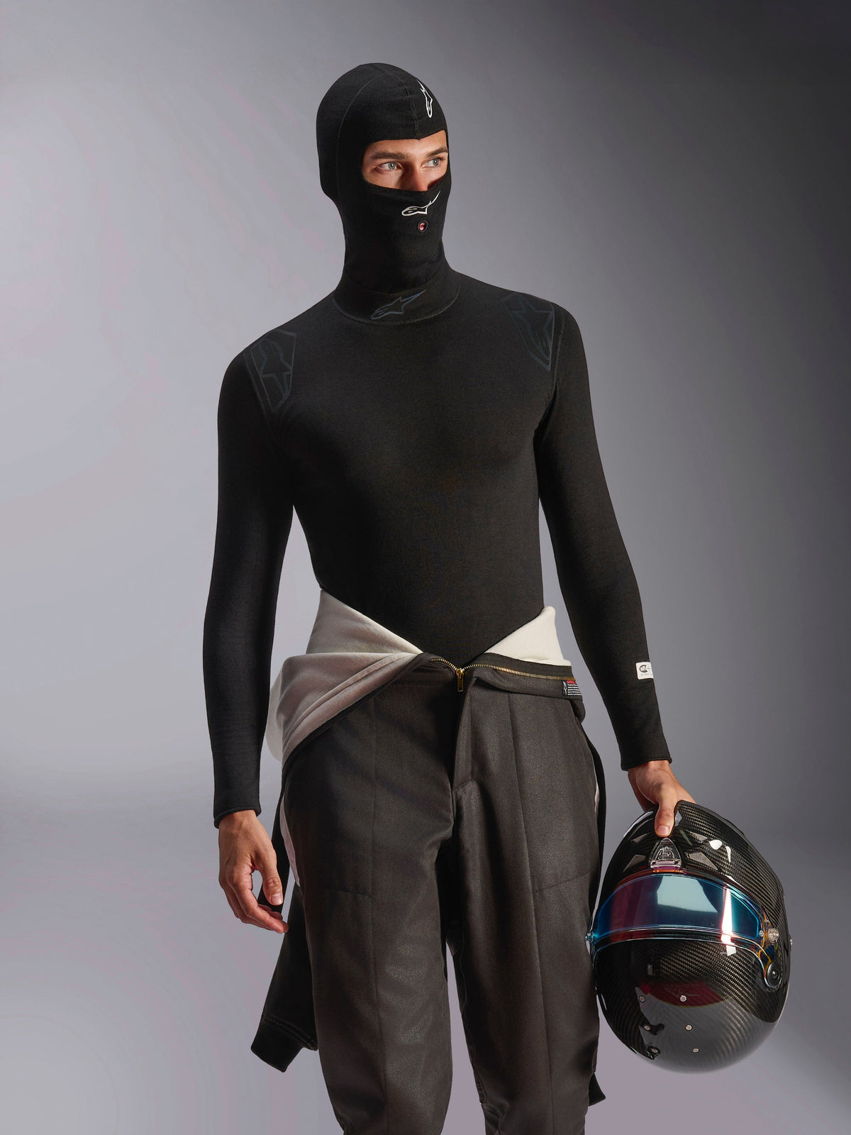 Mannelijk model dat de Alpinestars ZX Evo V3 bivakmuts in het zwart draagt, met witte logo's op het voorhoofd en de kin, gecombineerd met een zwarte basislaag met lange mouwen en een racehelm van koolstofvezel, professionele vlamvertragende motorsportuitrusting voor autoracen.