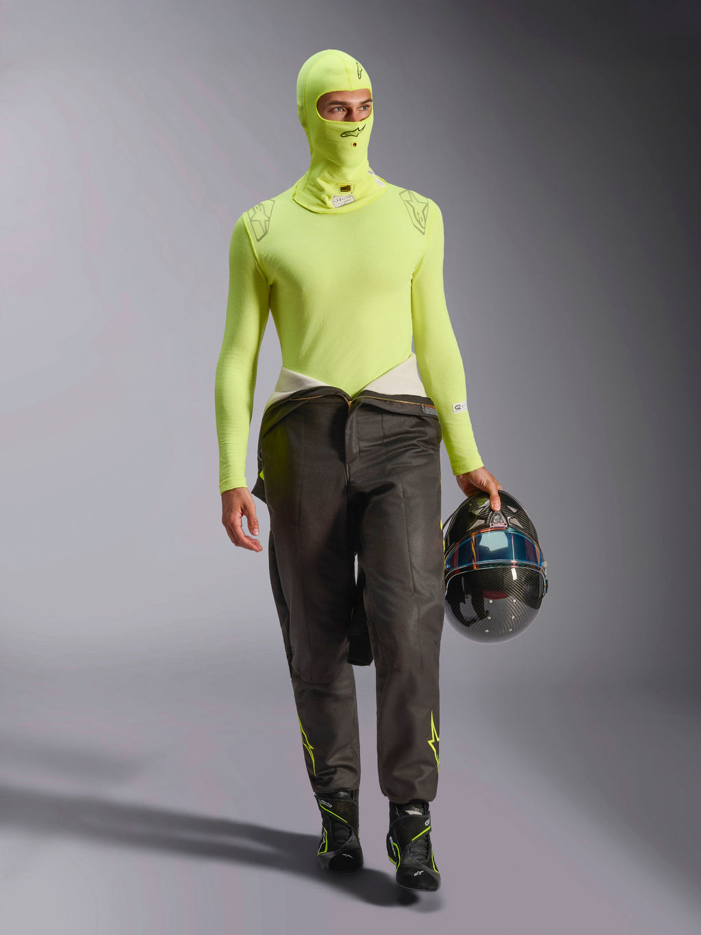 Mannelijke autocoureur in Alpinestars ZX Evo V3 bivakmuts in Yellow Fluo, bijpassende baselayer top met lange mouwen, donkere racebroek en zwarte racelaarzen, houdt een carbon fiber helm vast, professionele autorace-uitrusting