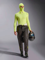 Mannelijke autocoureur in Alpinestars ZX Evo V3 bivakmuts in Yellow Fluo, bijpassende baselayer top met lange mouwen, donkere racebroek en zwarte racelaarzen, houdt een carbon fiber helm vast, professionele autorace-uitrusting
