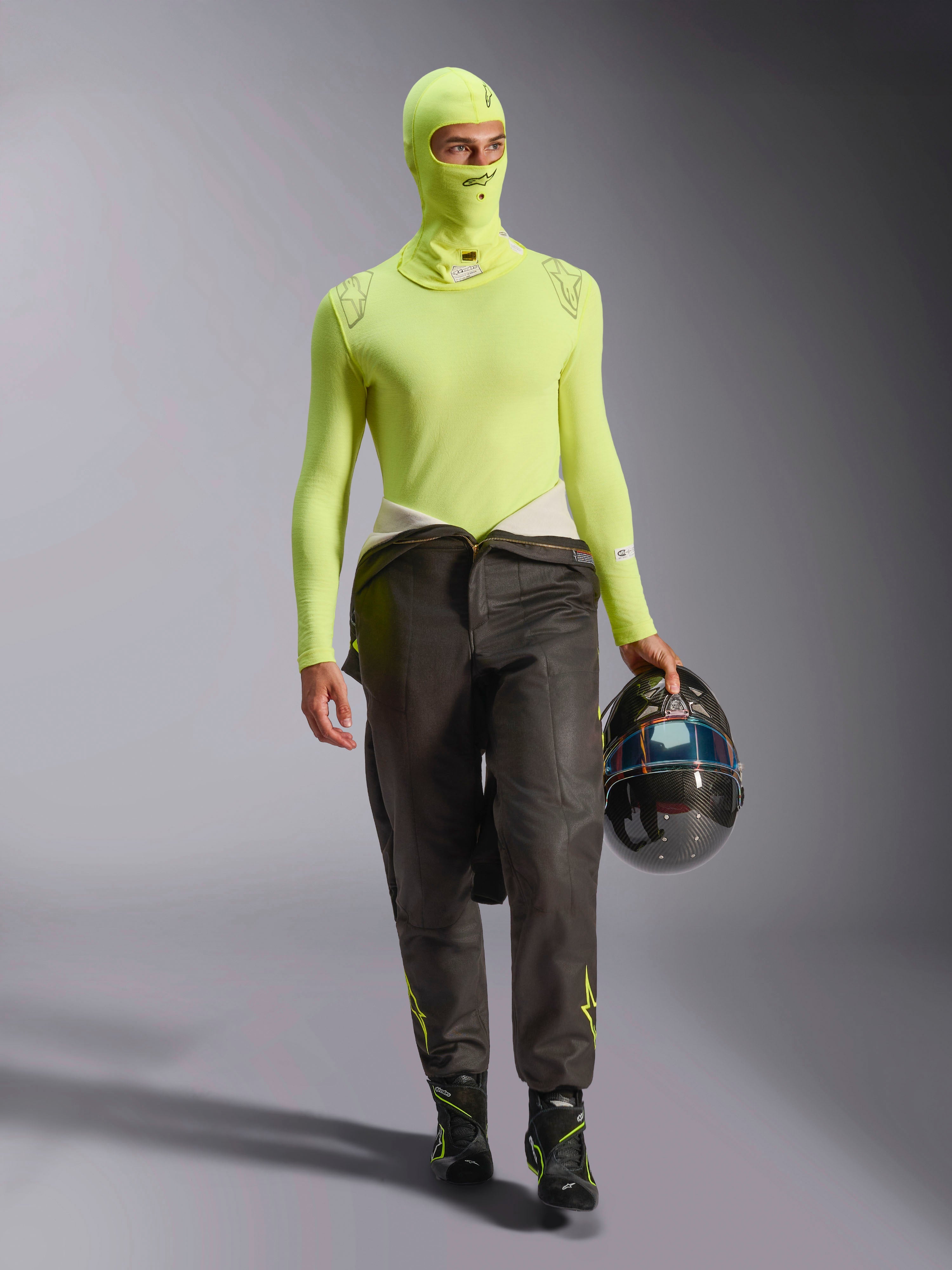 Mannelijke autocoureur in Alpinestars ZX Evo V3 bivakmuts in Yellow Fluo, bijpassende baselayer top met lange mouwen, donkere racebroek en zwarte racelaarzen, houdt een carbon fiber helm vast, professionele autorace-uitrusting