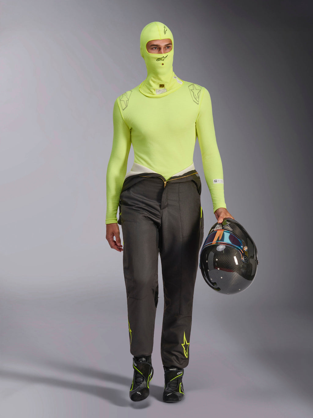 Alpinestars ZX Evo V3 Bivakmuts in Yellow Fluo, gedragen met bijpassende basislaag met lange mouwen en donkergrijze racebroek, mannelijk model houdt een helm van koolstofvezel vast, professionele autorace-uitrusting