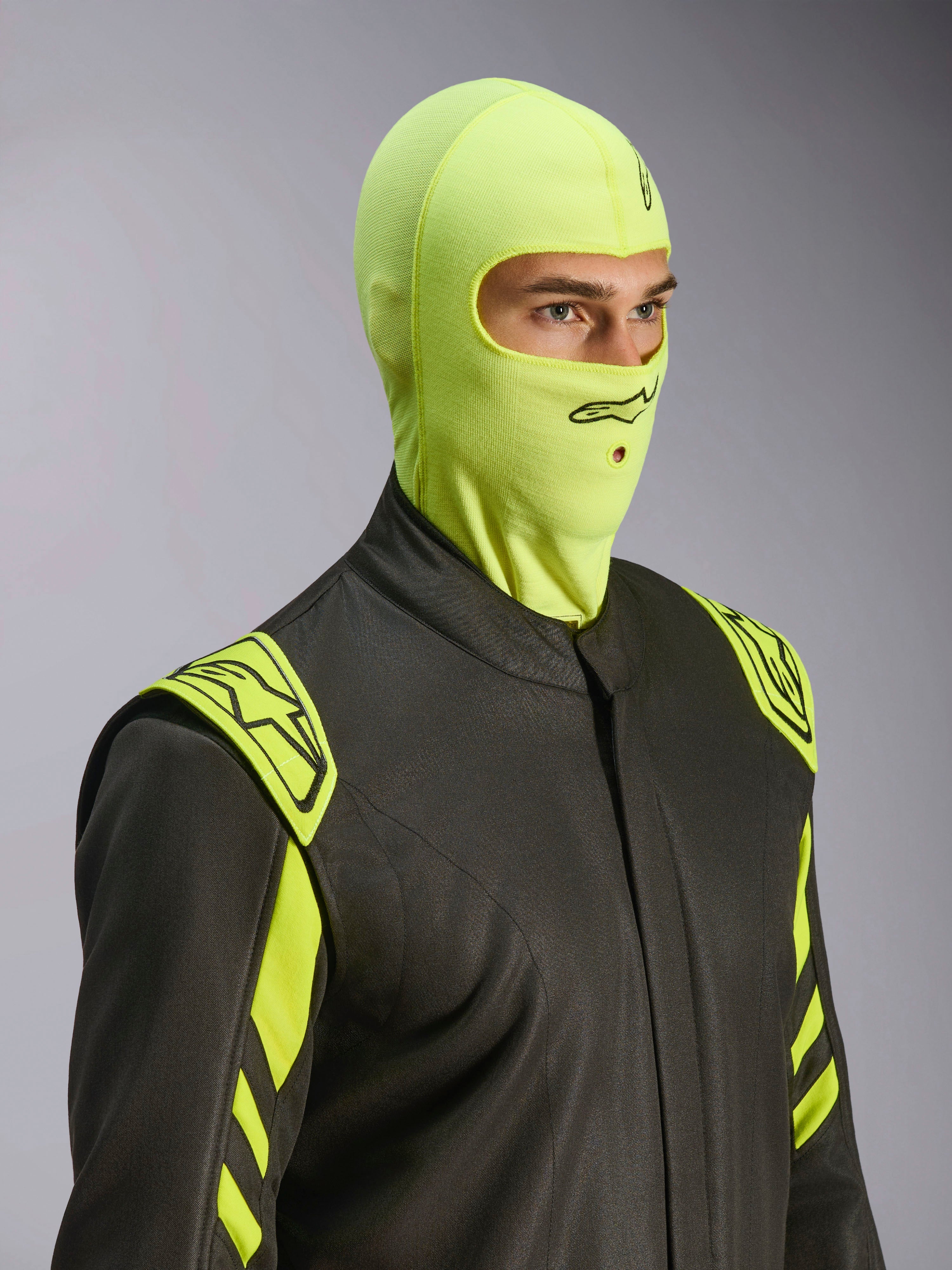 Mannelijk model draagt Alpinestars ZX Evo V3 bivakmuts in Yellow Fluo, vuurbestendige race-bivakmuts, gecombineerd met een donkergrijs autoracepak met neongele accenten, voorzien van zwart Alpinestars-logo en mondopening voor hydratatieslang, professionele beschermende autosportuitrusting