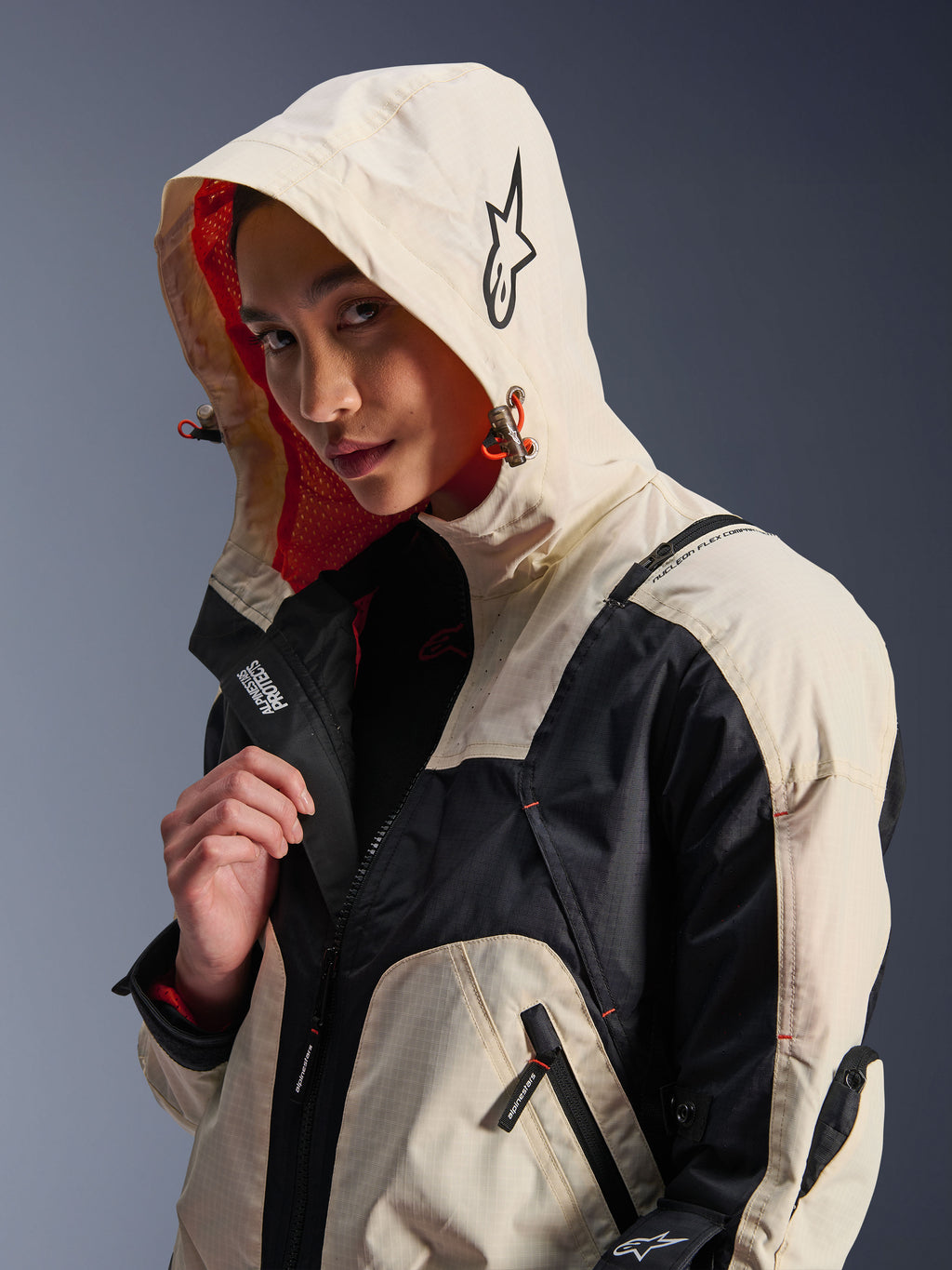 Alpinestars Molly Air Jacket in Black Almond, gedragen door een vrouwelijk model met de capuchon op, met crème en zwarte kleurvlakken, een opvallende oranje mesh voering en technische urban designelementen waaronder een sterlogo en ritsdetails.
