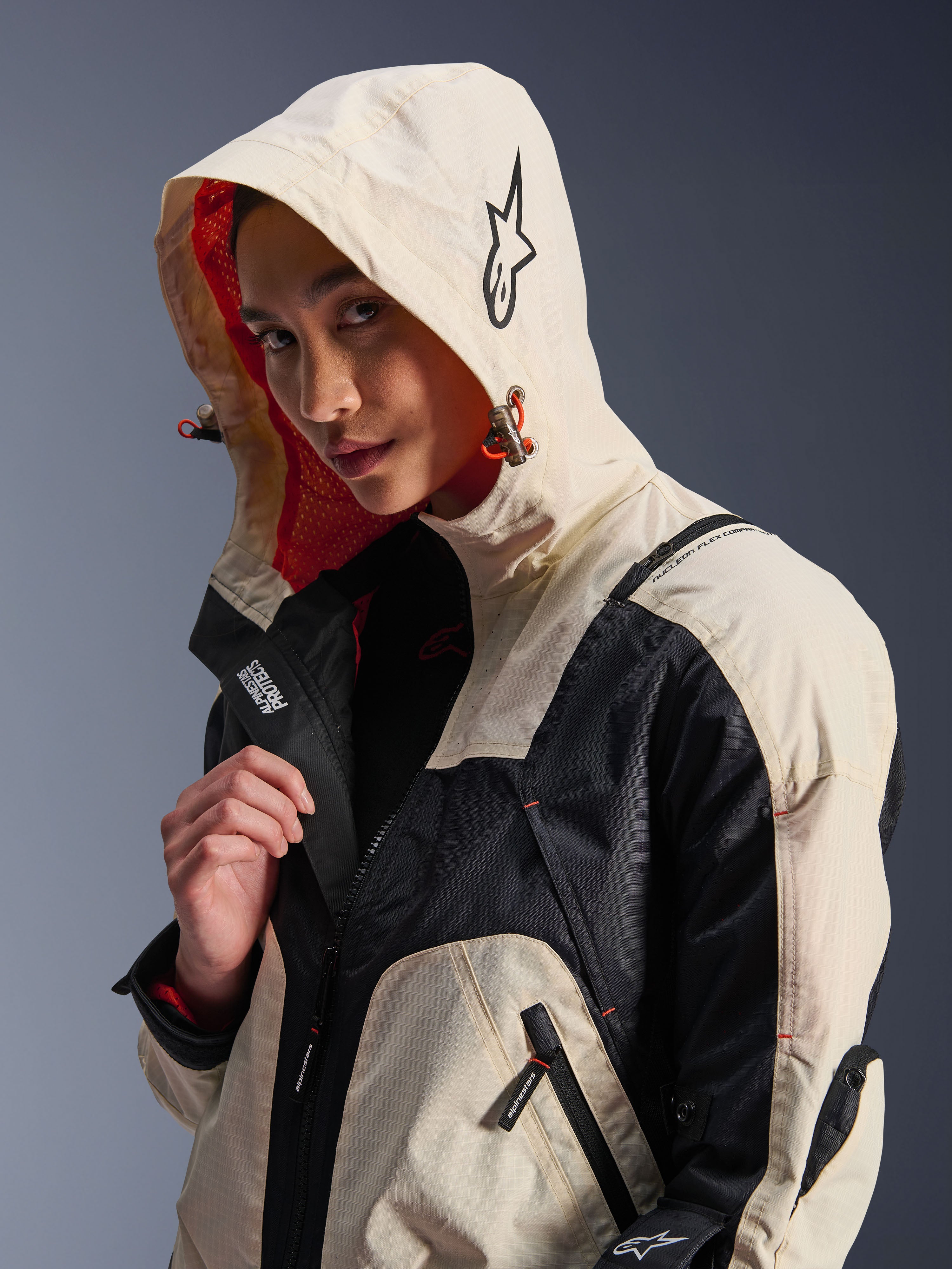 Alpinestars Molly Air Jacket in Black Almond, gedragen door een vrouwelijk model met de capuchon op, met crème en zwarte kleurvlakken, een opvallende oranje mesh voering en technische urban designelementen waaronder een sterlogo en ritsdetails.