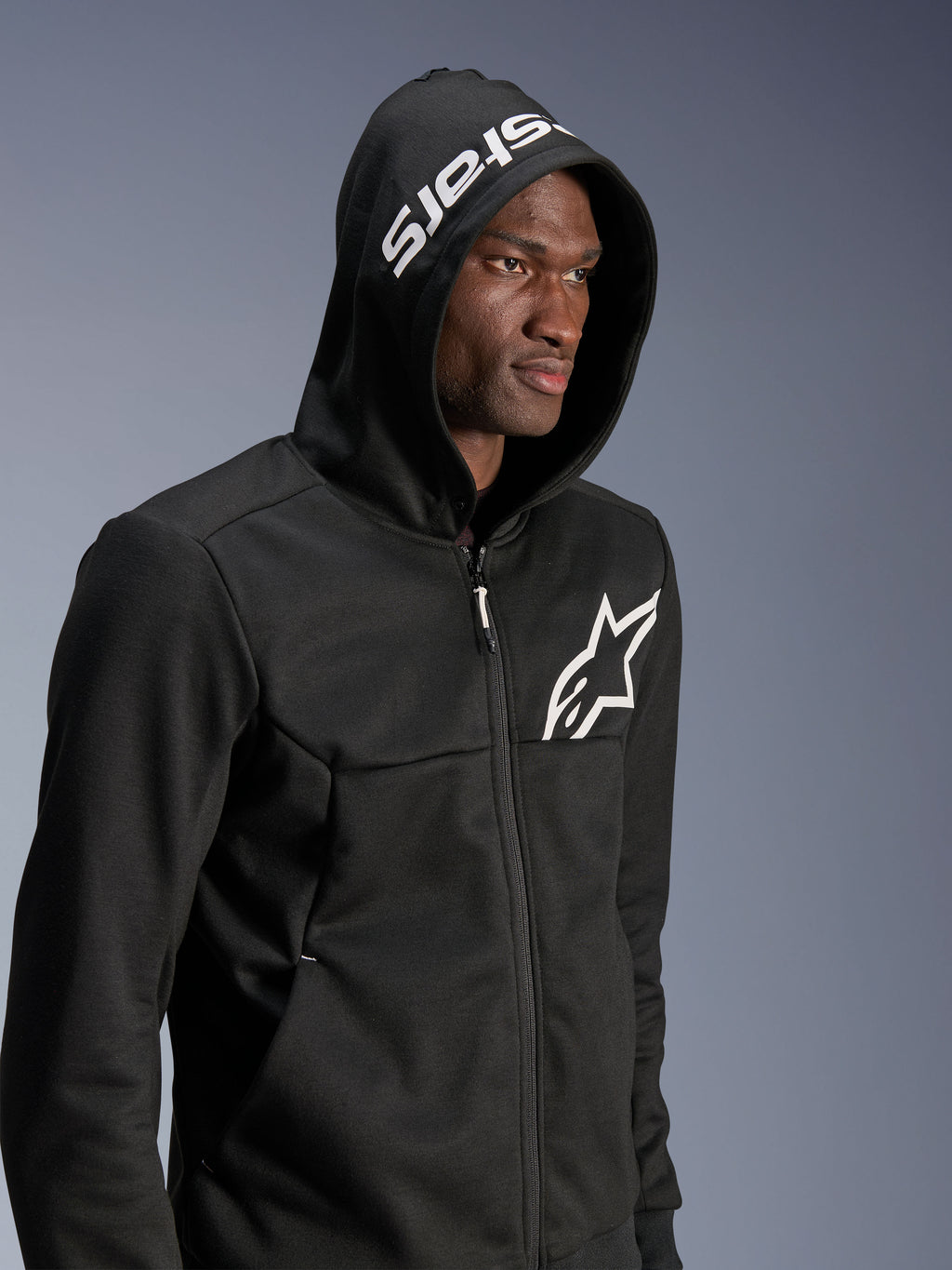 Alpinestars Chrome V2 Sport Hoodie, sweat à capuche de conduite urbaine noir porté par un mannequin avec la capuche relevée, logo étoile blanc sur la poitrine et marquage blanc sur la capuche, construction en polaire technique, style streetwear moto décontracté
