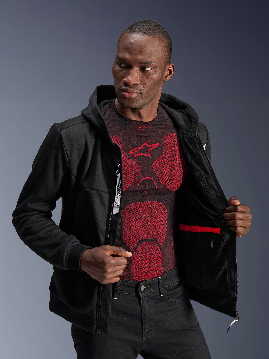 Alpinestars Chrome V2 Sport Hoodie, noir, mannequin homme portant le sweat à capuche ouvert pour montrer la doublure intérieure, la poche étanche rouge et le marquage interne, porté sur une couche de base rouge et noire, équipement de moto urbain décontracté