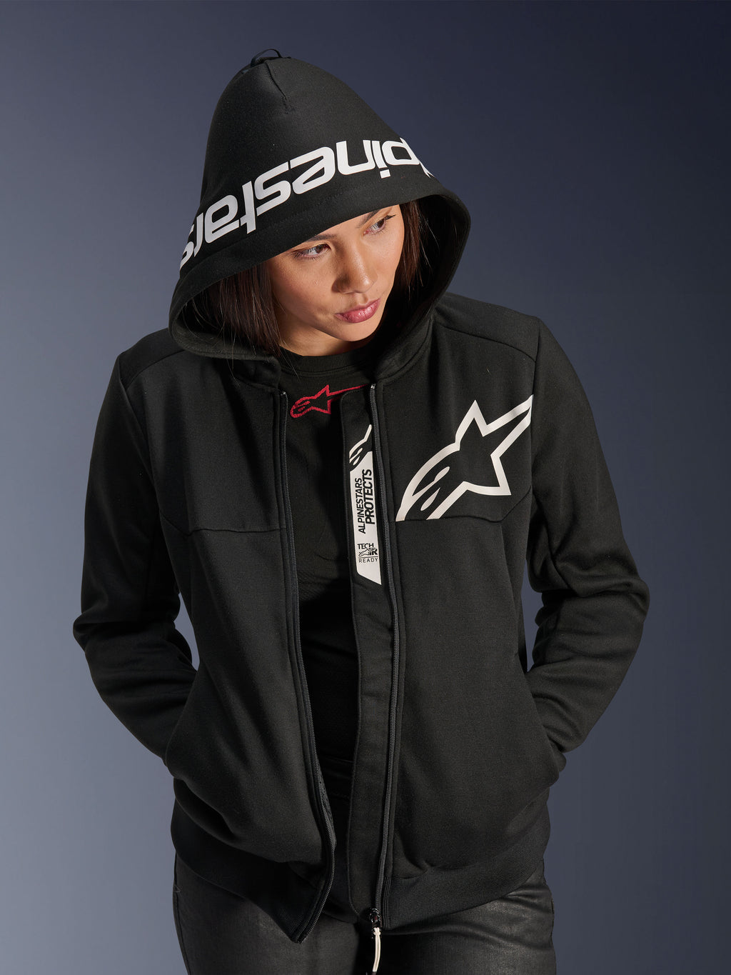 Alpinestars Stella Chrome V2 Sport Hoodie pour femme, noir et blanc, sweat à capuche de moto avec un logo étoile blanche sur la poitrine et le marquage Alpinestars sur le bord de la capuche, femme portant la capuche, style sport urbain décontracté avec détails compatibles Tech-Air.