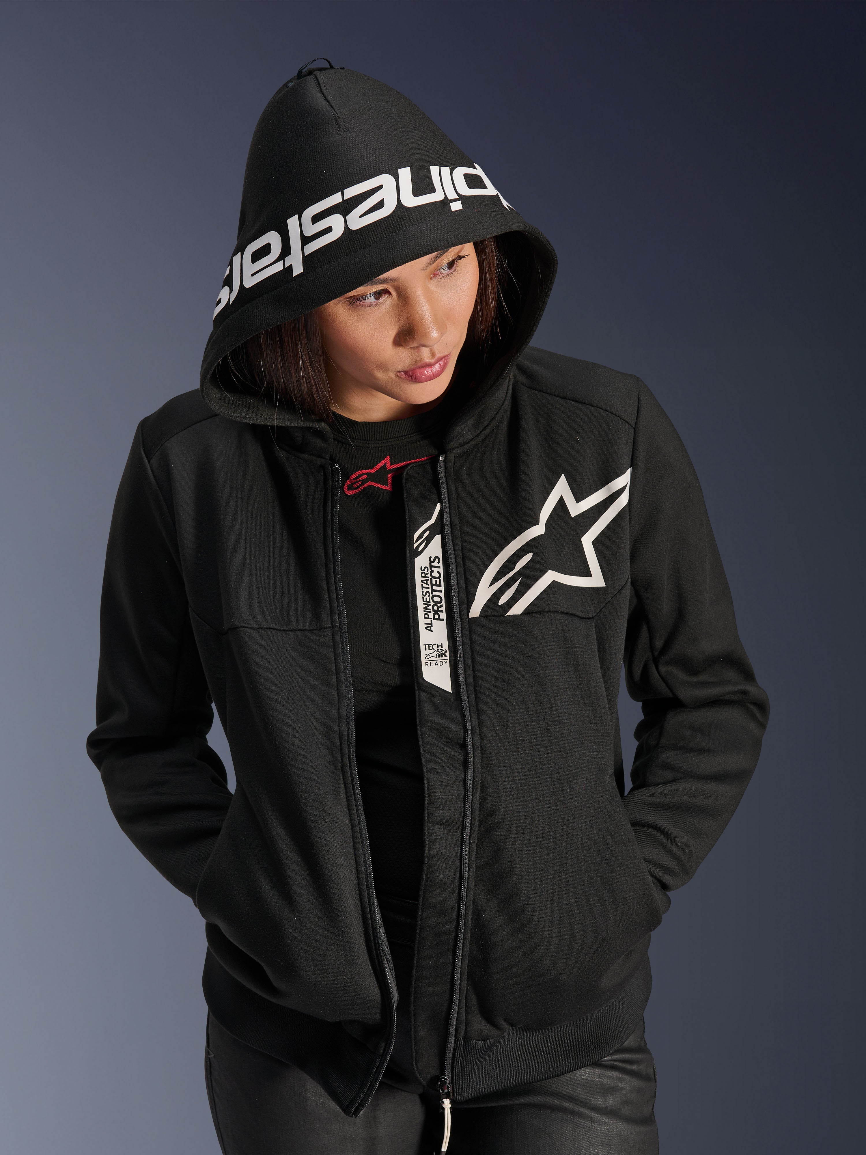 Alpinestars Stella Chrome V2 Sport Hoodie pour femme, noir et blanc, sweat à capuche de moto avec un logo étoile blanche sur la poitrine et le marquage Alpinestars sur le bord de la capuche, femme portant la capuche, style sport urbain décontracté avec détails compatibles Tech-Air.