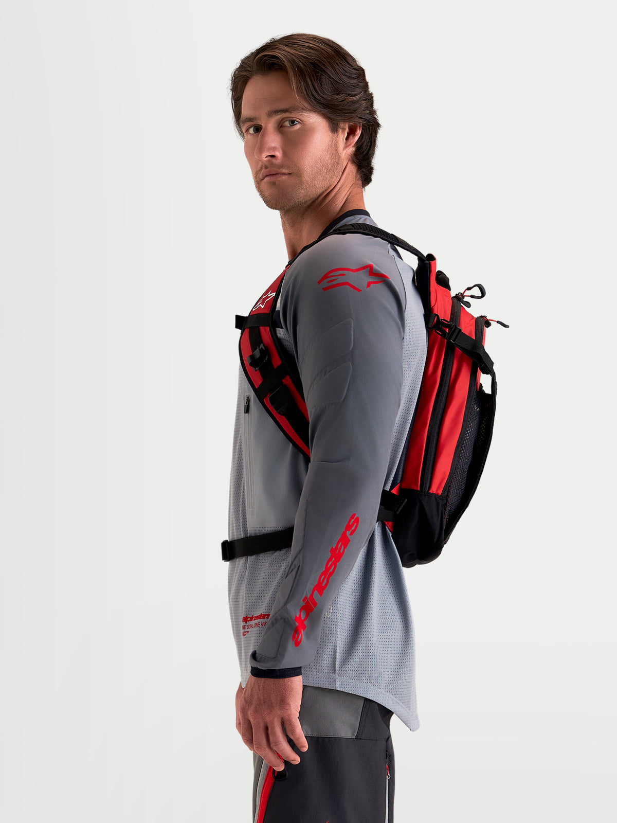 Alpinestars AT-9 Rucksack, Motocross-Rucksack in Schwarz und Leuchtrot, Seitenprofil-Ansicht getragen von einem männlichen Model mit passender MX-Ausrüstung, leichte Textilkonstruktion und ergonomisches Tragesystem