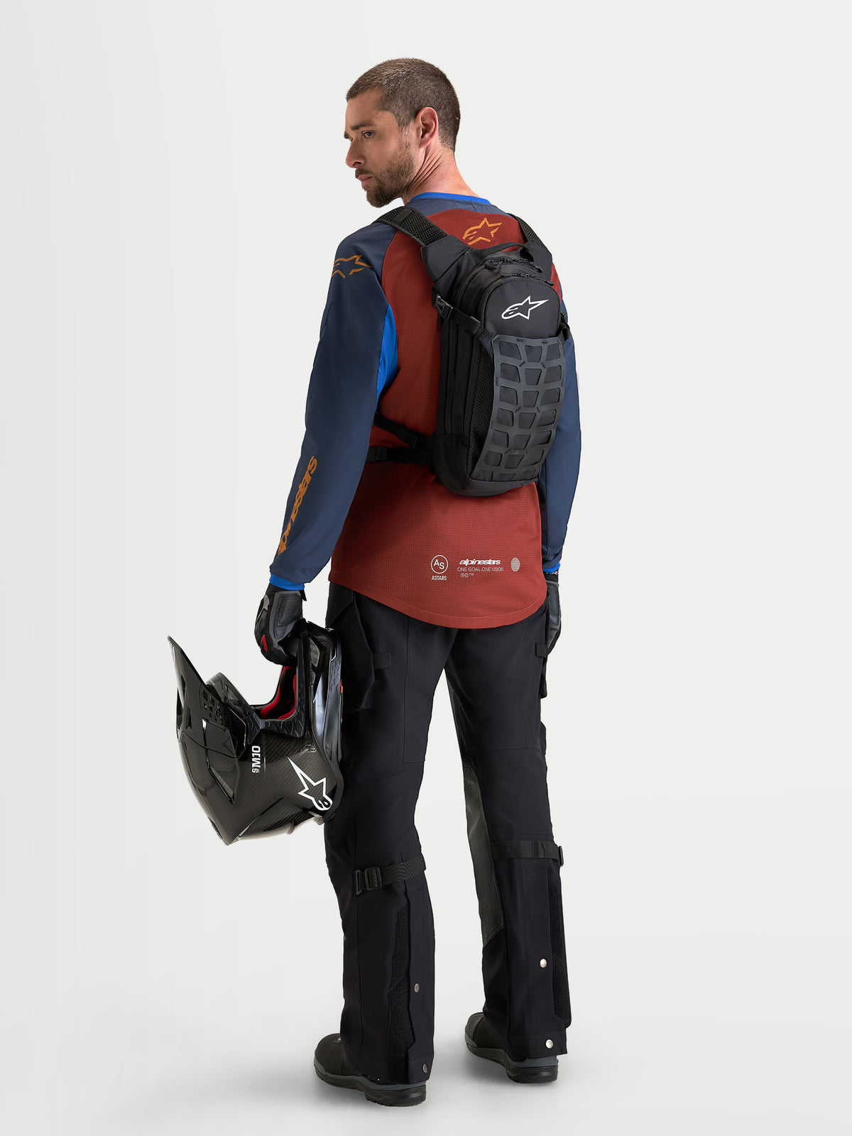Alpinestars Sac À Dos AT-9, sac à dos de motocross noir et gris foncé, porté par un modèle de dos tenant un casque et portant un équipement de moto assorti