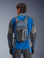 Alpinestars AT-15 Rucksack, Adventure-Motorradrucksack, Dunkelgrau und Tiefgrau, getragen von einem Fahrer über einer blau-grauen Jacke, mit All-Terra-Branding, ergonomischem strukturellem Design, verstellbaren Riemen und blauen Reißverschluss-Akzenten.