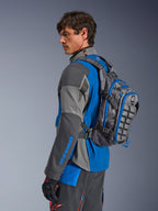 Alpinestars AT-15 Rucksack, Motorrad-Adventure-Rucksack, Dunkelgrau mit blauen Akzenten, getragen von einem Fahrer in farblich abgestimmter grau-blauer technischer Fahrbekleidung, mit MOLLE-Panel und robuster Konstruktion für Offroad-Abenteuer.