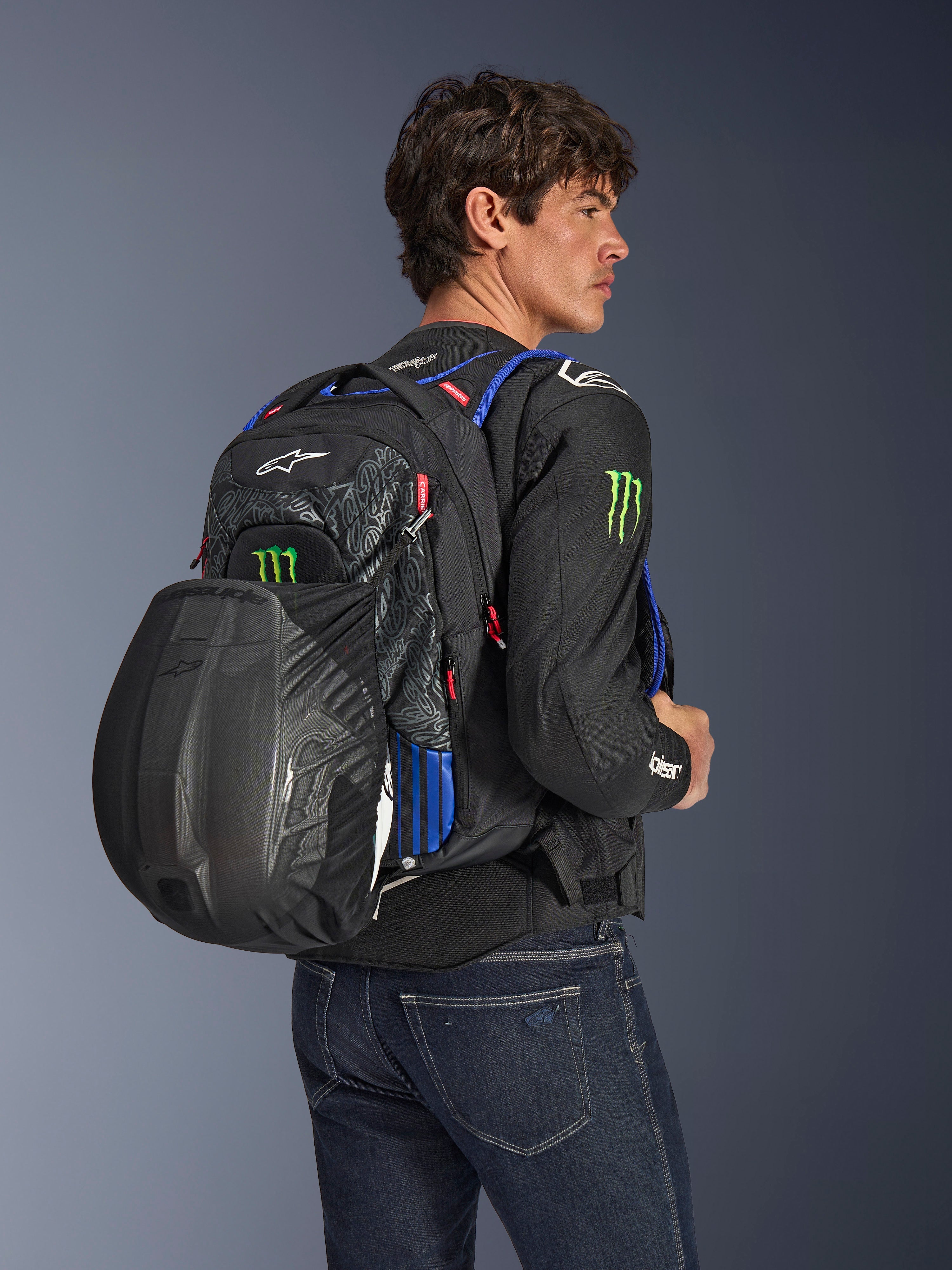 Alpinestars FQ20 City Hunter V2 Monster rugzak, stedelijke motorrugzak, zwart met fluo rode, blauwe en groene accenten, model draagt een zwarte helm in de mesh-houder aan de achterzijde, gestyled met een zwarte motorjas en blauwe jeans.