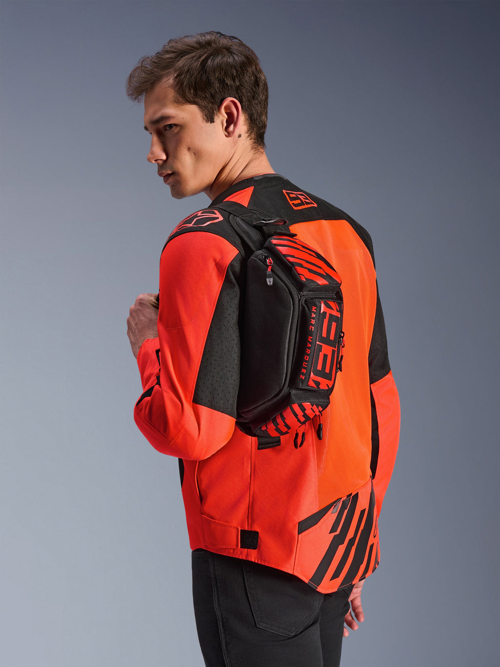 Alpinestars MM93 Waist Bag, heuptas, zwart met Red Fluo en Bright Red, door een model over de rug gedragen over een rood-zwarte motorjas, voorzien van opvallende MM93-branding en Marc Marquez-graphics