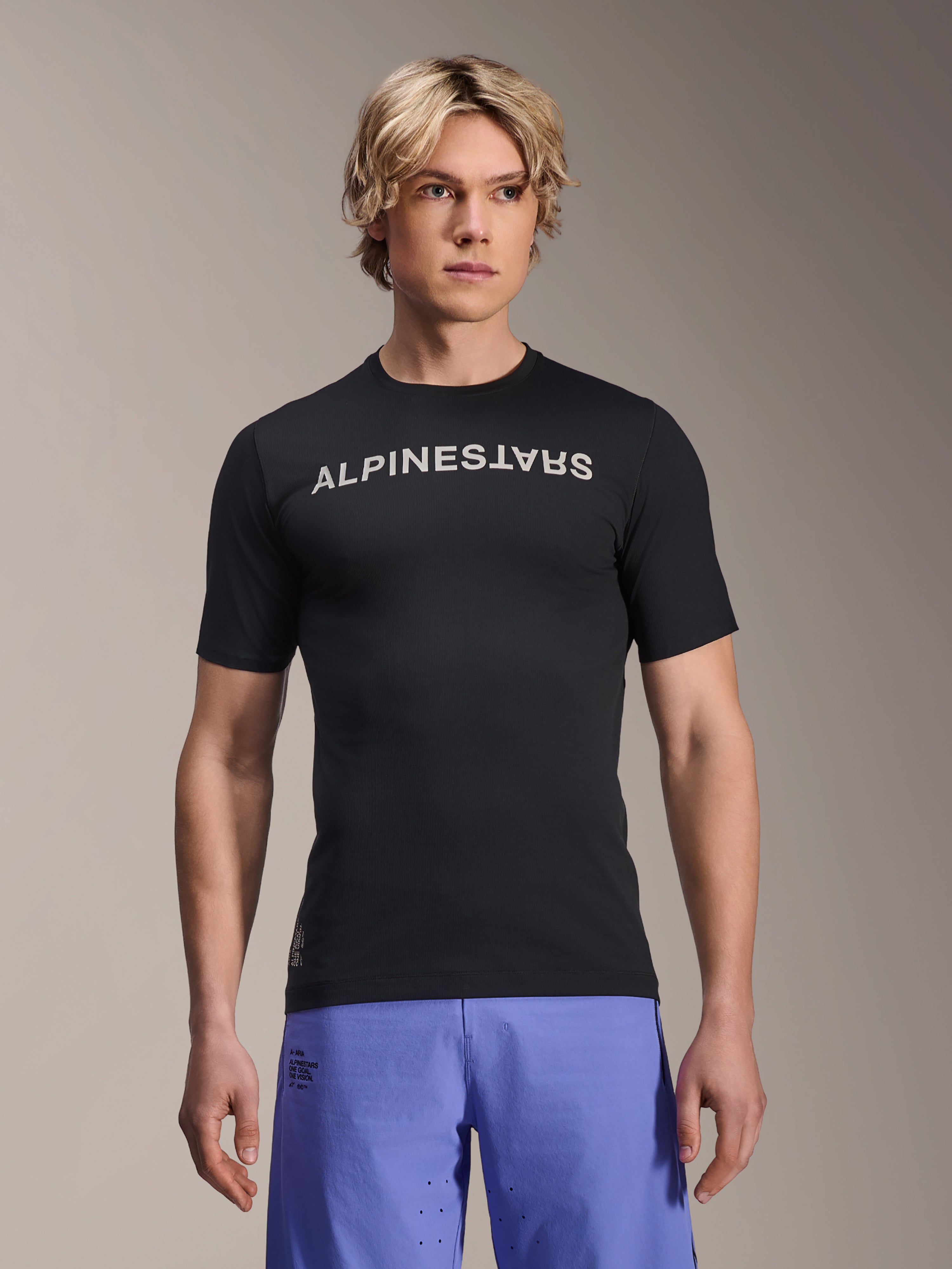 Alpinestars A-Aria Switch Jersey, Short Sleeve MTB Jersey, Black with white Alpinestars wordmark on chest, worn by male model with purple shorts, lightweight performance fabric for trail riding