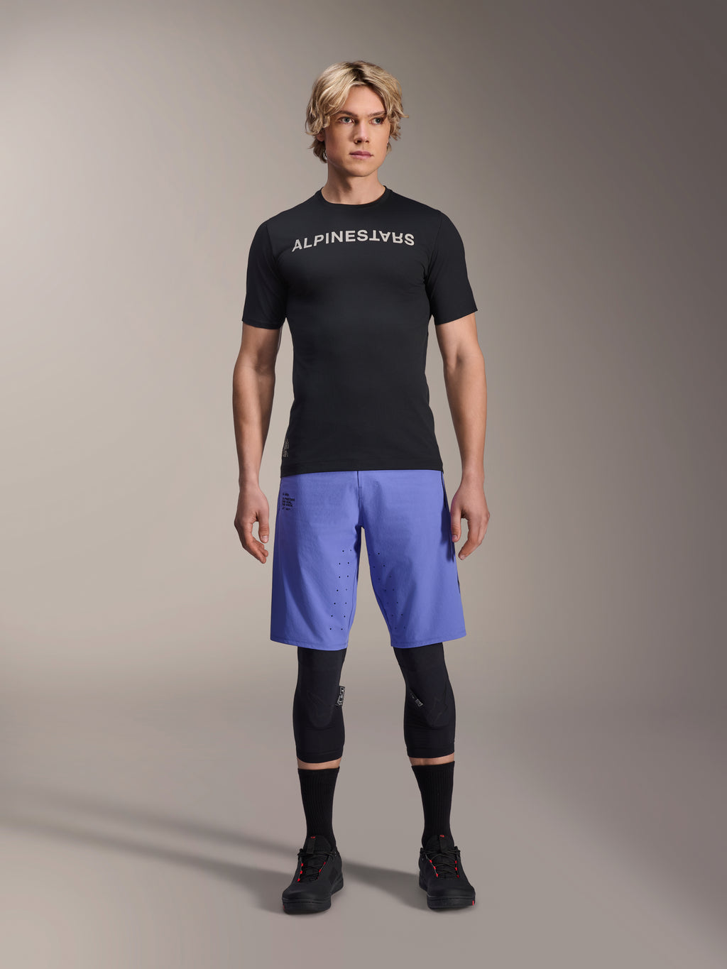 Maillot Alpinestars A-Aria Switch, maillot de VTT à manches courtes, noir avec logo Alpinestars blanc en miroir, porté par un mannequin homme avec un short de cyclisme bleu et des genouillères noires, vêtement de performance respirant pour le trail