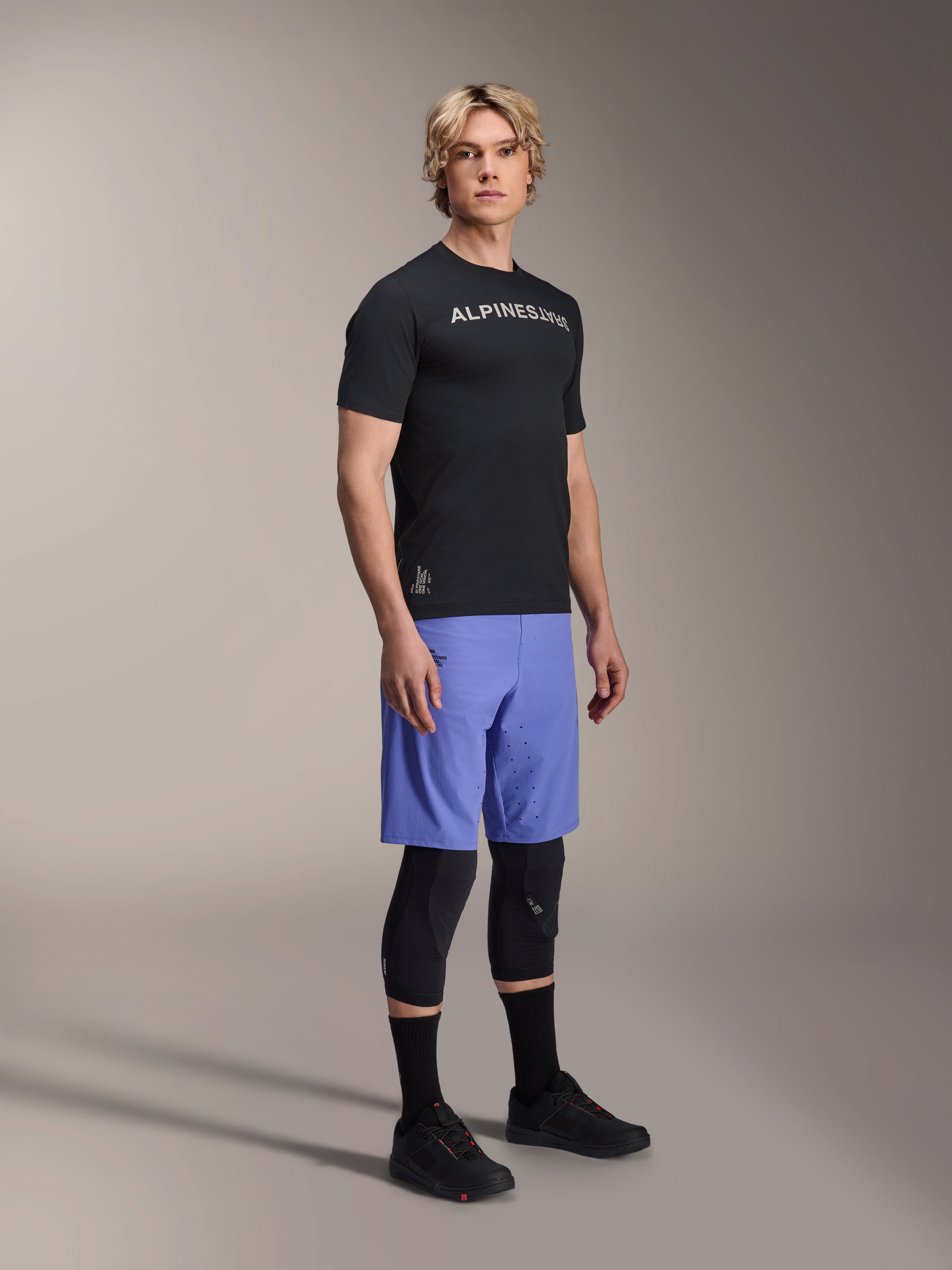 Mannequin homme portant le maillot à manches courtes Alpinestars A-Aria Switch noir avec logo blanc sur la poitrine, associé à un short de VTT bleu clair, des genouillères noires et des chaussures de cyclisme noires, prise de vue en studio en pied pour équipement de VTT haute performance