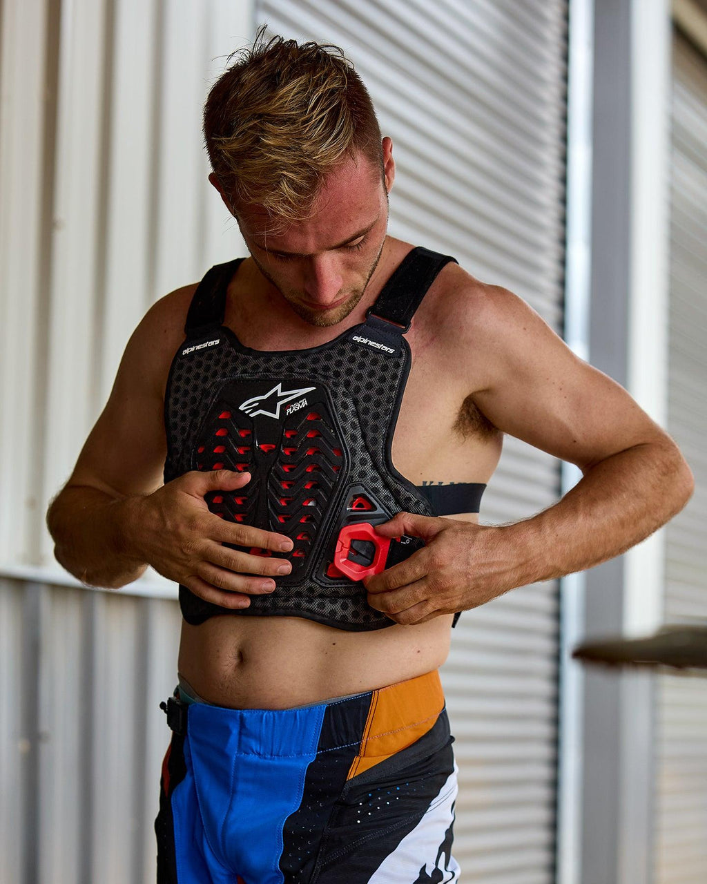 Man die de Alpinestars A-5 Plasma borstbeschermer aantrekt, zwart met rode accenten en wit logo, voorzien van een honingraatstructuur, gedragen met blauwe en oranje motorcrossbroek, voorbereidende setting voor off-road racen