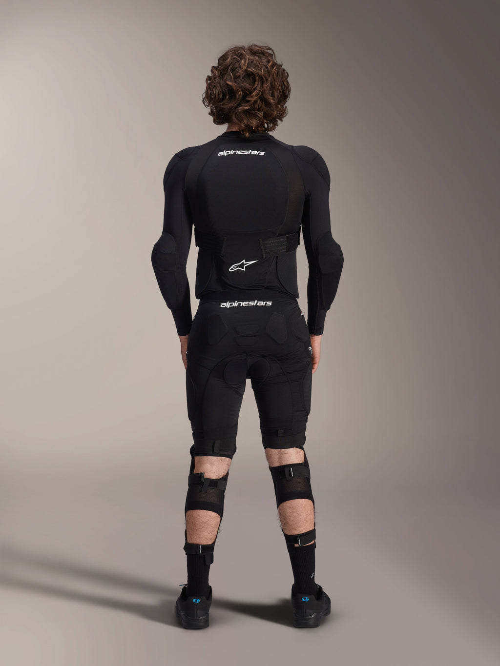 Veste de protection Alpinestars A-Impact Plasma - Manches longues, vue de dos, noire avec logos Alpinestars blancs, portée avec un short de protection et des genouillères pour une protection intégrale en VTT