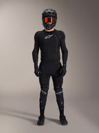 Cycliste de VTT portant un équipement de protection complet, veste de protection Alpinestars A-Impact Plasma en noir et blanc, logo blanc sur la poitrine et tissu en maille, avec un casque noir, des lunettes teintées de rouge et des genouillères, ensemble de protection VTT haute performance coordonné.