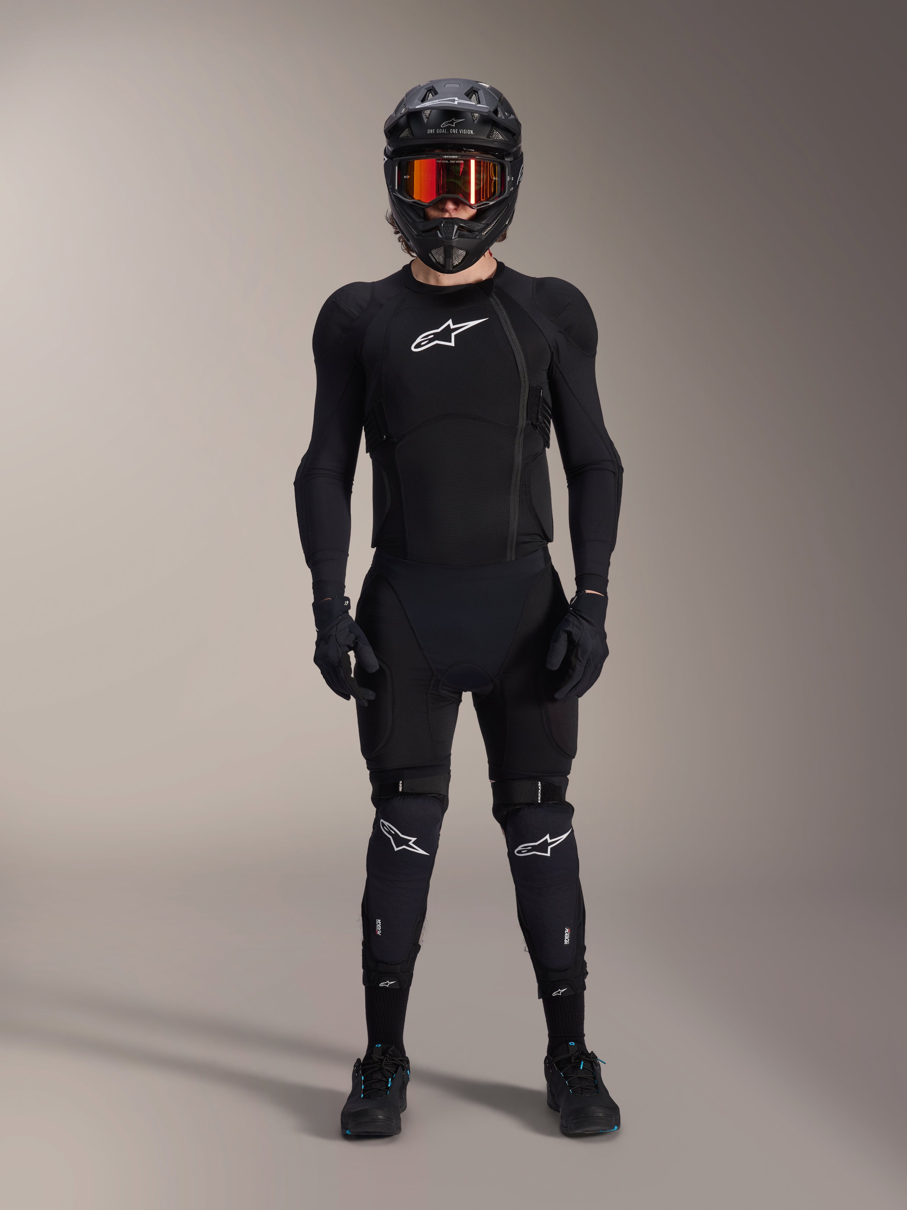 Cycliste de VTT portant un équipement de protection complet, veste de protection Alpinestars A-Impact Plasma en noir et blanc, logo blanc sur la poitrine et tissu en maille, avec un casque noir, des lunettes teintées de rouge et des genouillères, ensemble de protection VTT haute performance coordonné.