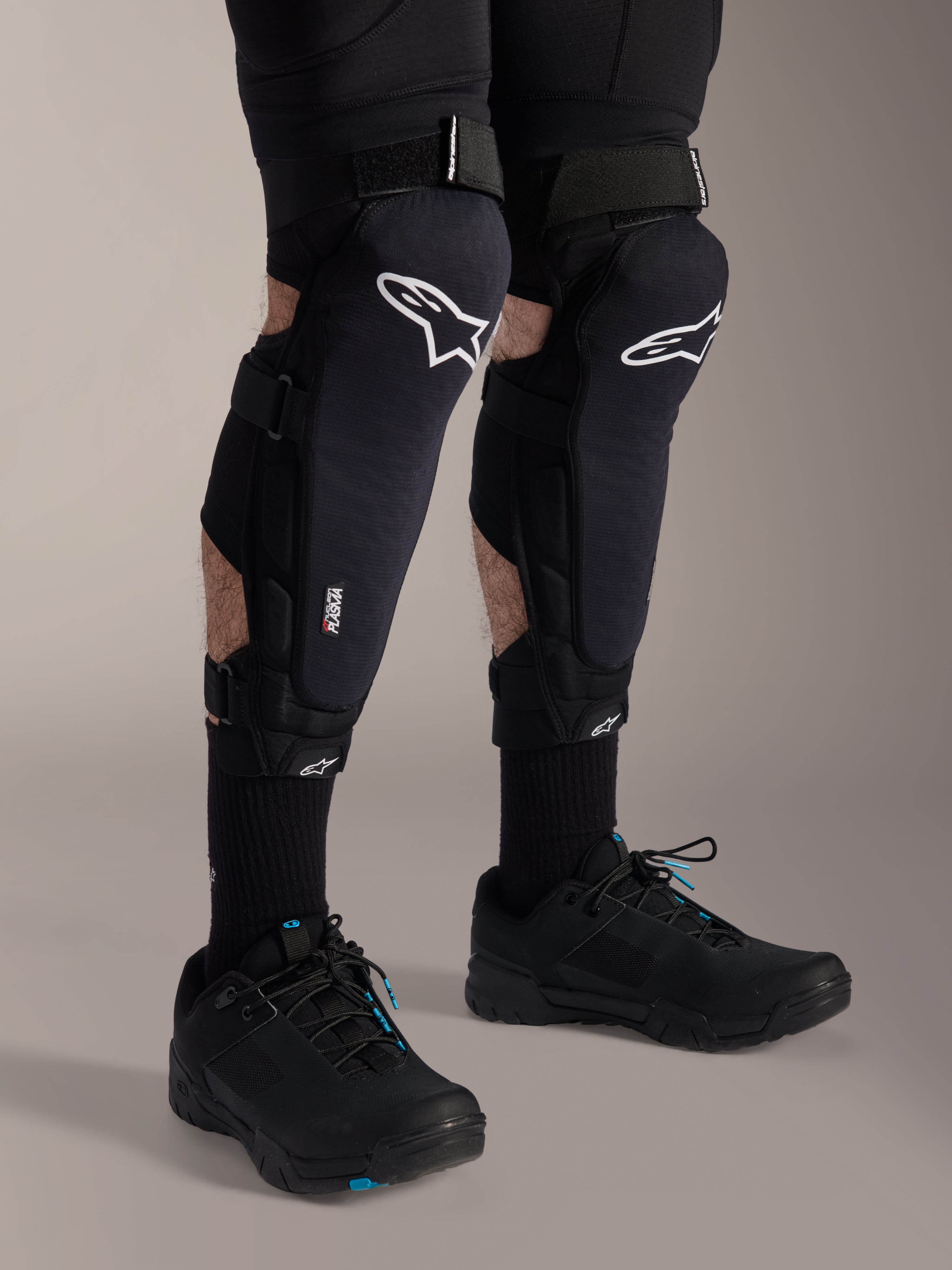 Protections genoux et tibias Alpinestars A-Impact Plasma Pro, noir et blanc, avec logo Alpinestars blanc et système de fermeture à triple sangle, portées avec chaussettes noires et chaussures de sport noires, protection durable pour le cyclisme tout-terrain