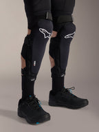 Alpinestars A-Impact Plasma Pro kniebeschermers, zwart en wit, met Nucleon PLASMA-technologie branding, gedragen met zwarte sokken en zwarte mountainbikeschoenen, stevige klittenbandsluitingen, ergonomische beschermingsuitrusting voor off-road fietsen