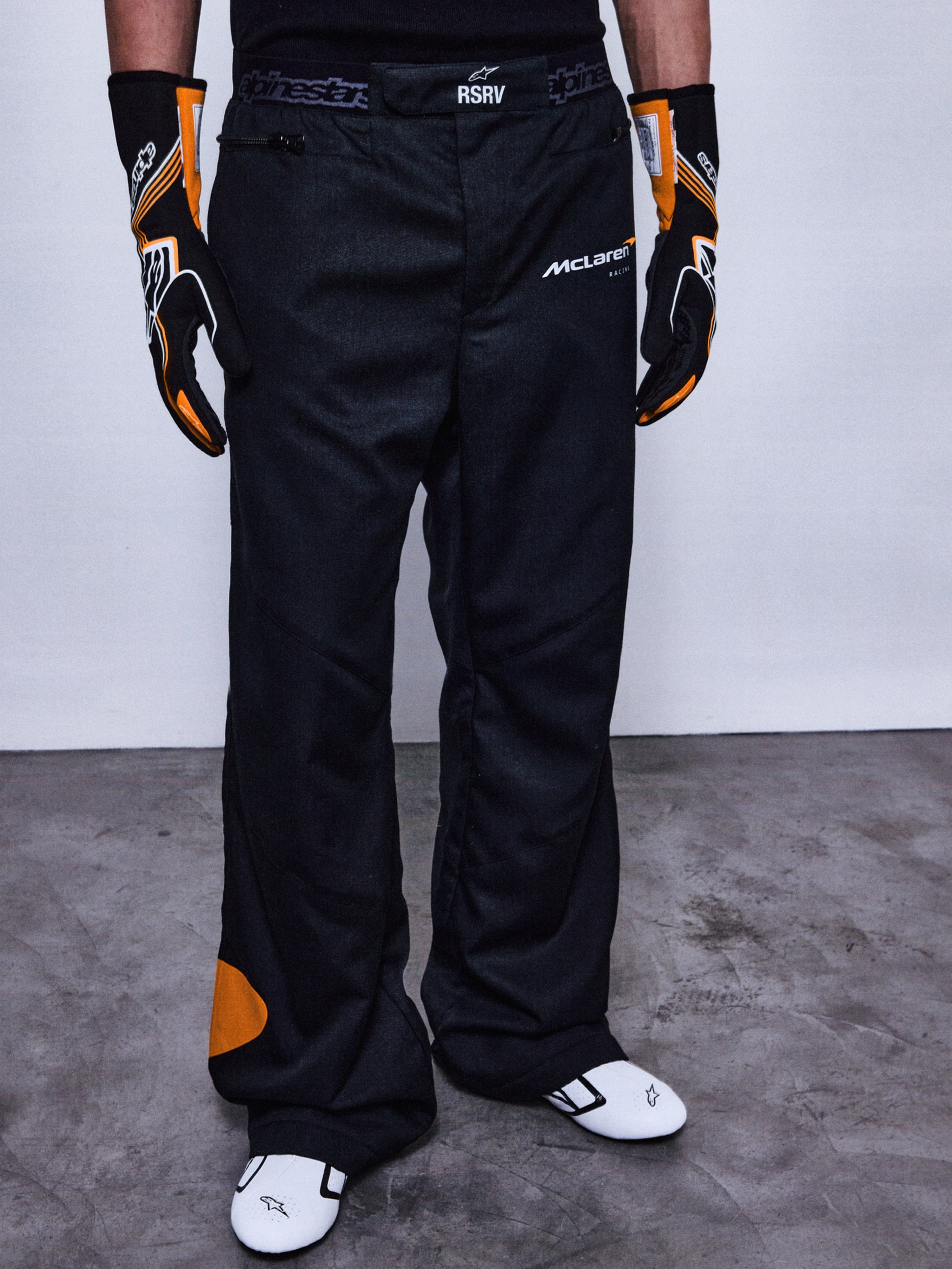 Alpinestars RSRV Arena Suit Pants, pantalon de course noir avec logos McLaren et Alpinestars, détails orange, porté avec gants noirs et orange et chaussures de course blanches
