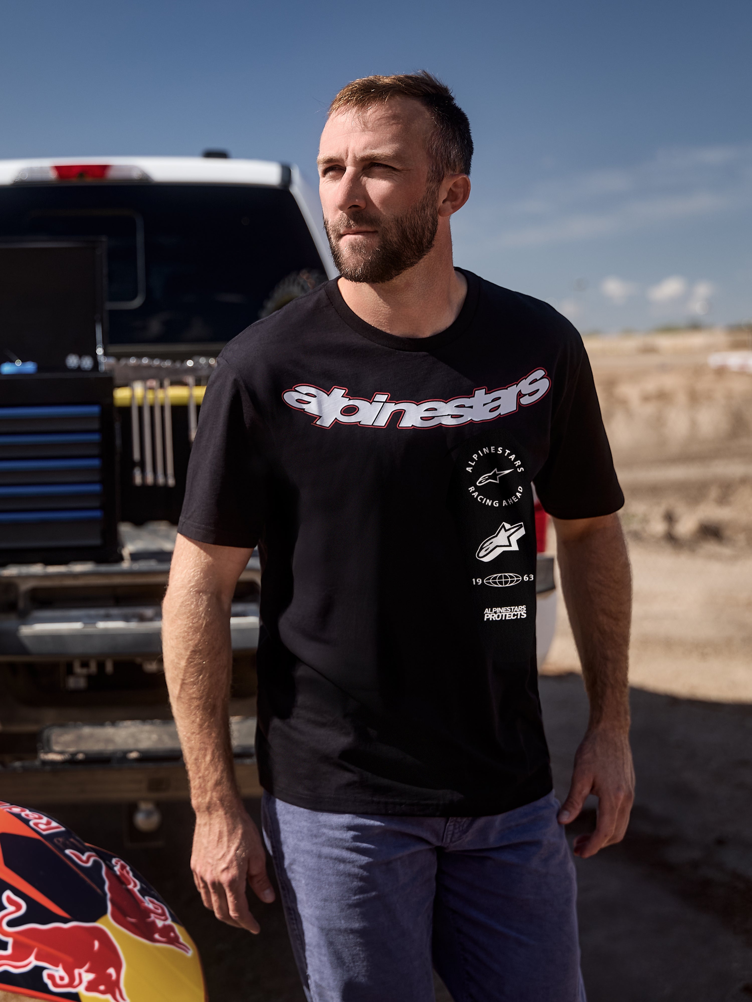 Alpinestars Abus CSF Tee, kurzärmeliges T-Shirt, Schwarz, mit einem großen gebogenen Alpinestars-Schriftzug und mehreren gestapelten Logos, getragen von einem Mann in einer Motorsport-Paddock-Umgebung, lässige Lifestyle-Streetwear
