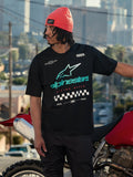 Alpinestars Aplomb Oversized CSF Tee, T-shirt met korte mouwen, zwart, met een groot blauwgroen en wit Astars-logo met kleurverloop en een geblokte vlag-graphic, gedragen met een koraalrode beanie, staand naast een rode motorfiets, casual lifestyle streetwear