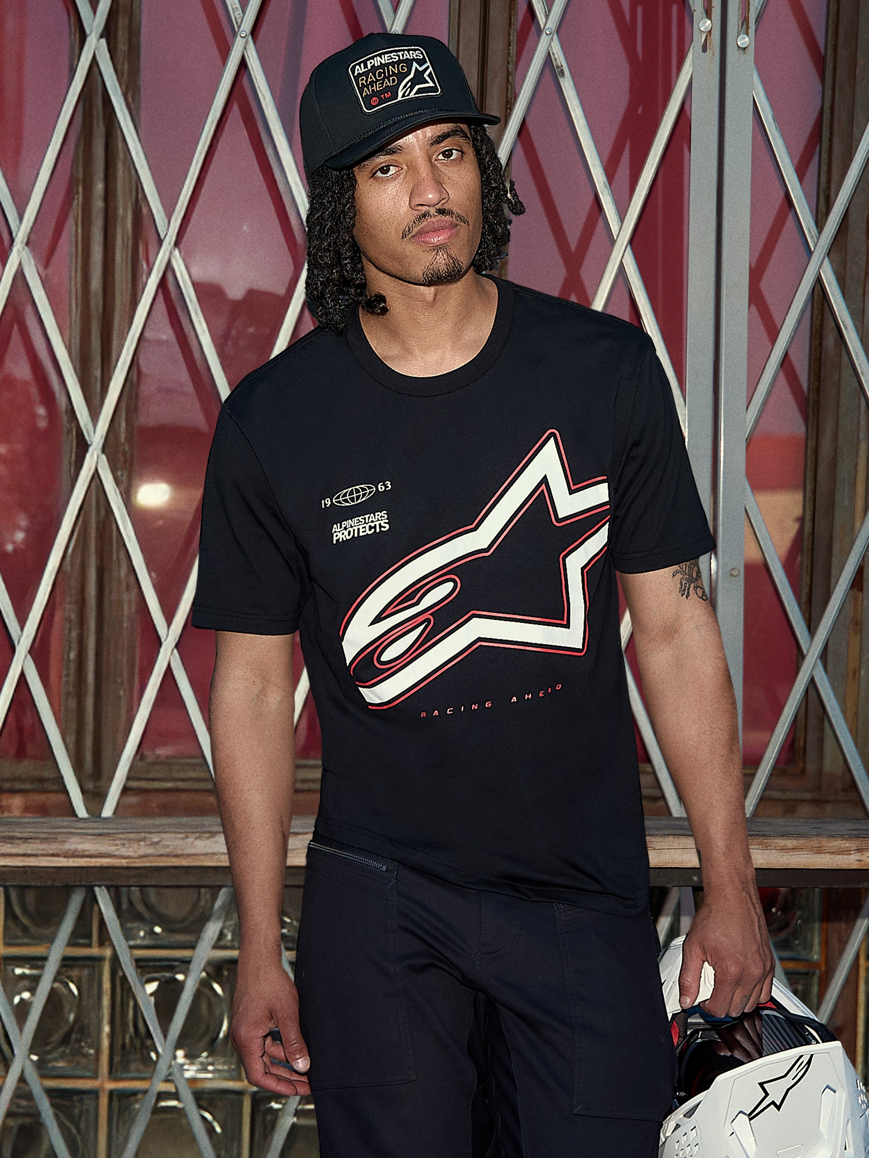 Alpinestars Authority CSF Tee, T-shirt met korte mouwen, zwart, groot wit en rood Astars outline-logo met 'Alpinestars Protects' graphic, lifestylefoto van een man met een zwarte truckerpet die een witte helm vasthoudt, casual motorsport streetwear