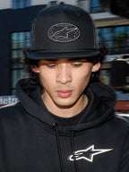 Alpinestars Contender Trucker pet, zwarte lifestyle pet met mesh achterkant, gestructureerde kroon voorzien van een geborduurde Astars logo patch, gedragen door een man met een zwarte Alpinestars hoodie, casual motorsport-geïnspireerde streetwear.