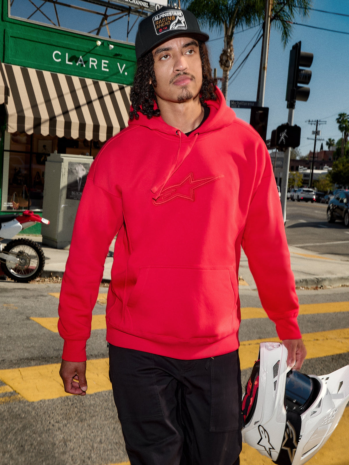 Alpinestars Emergent Oversized Hoodie, Rot, 3D-gesticktes Astars-Logo auf der Brust, getragen mit schwarzer Hose und Baseball-Cap, hält einen weißen Motocross-Helm, lässiger urbaner Streetwear-Stil