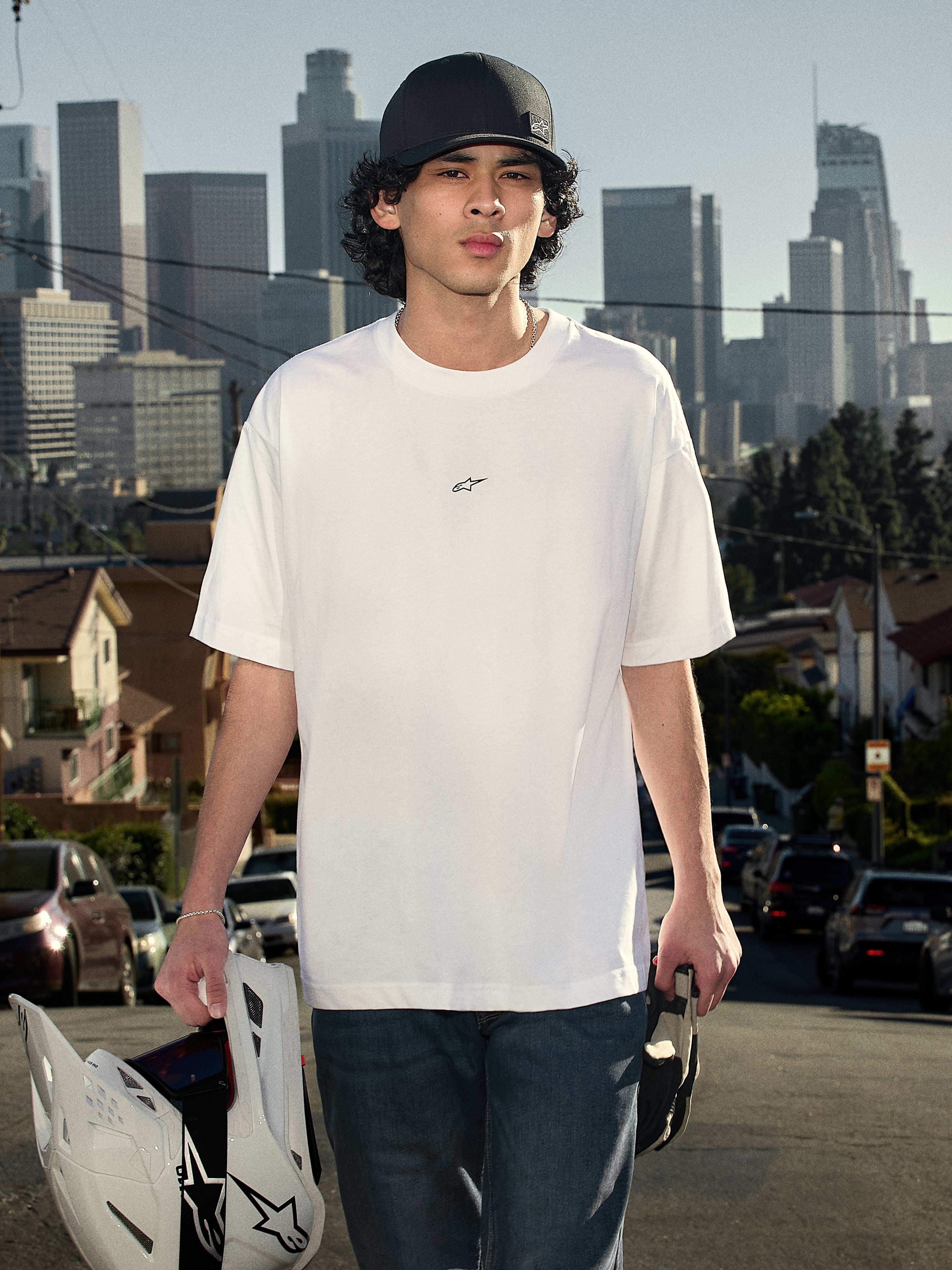 Alpinestars Focused Oversized CSF Tee, T-shirt met korte mouwen, wit, met een klein zwart Astars-logo op de borst, lifestyle-opname van een persoon die het shirt draagt en een motocrosshelm vasthoudt met een stads-skyline op de achtergrond, casual motorsport streetwear