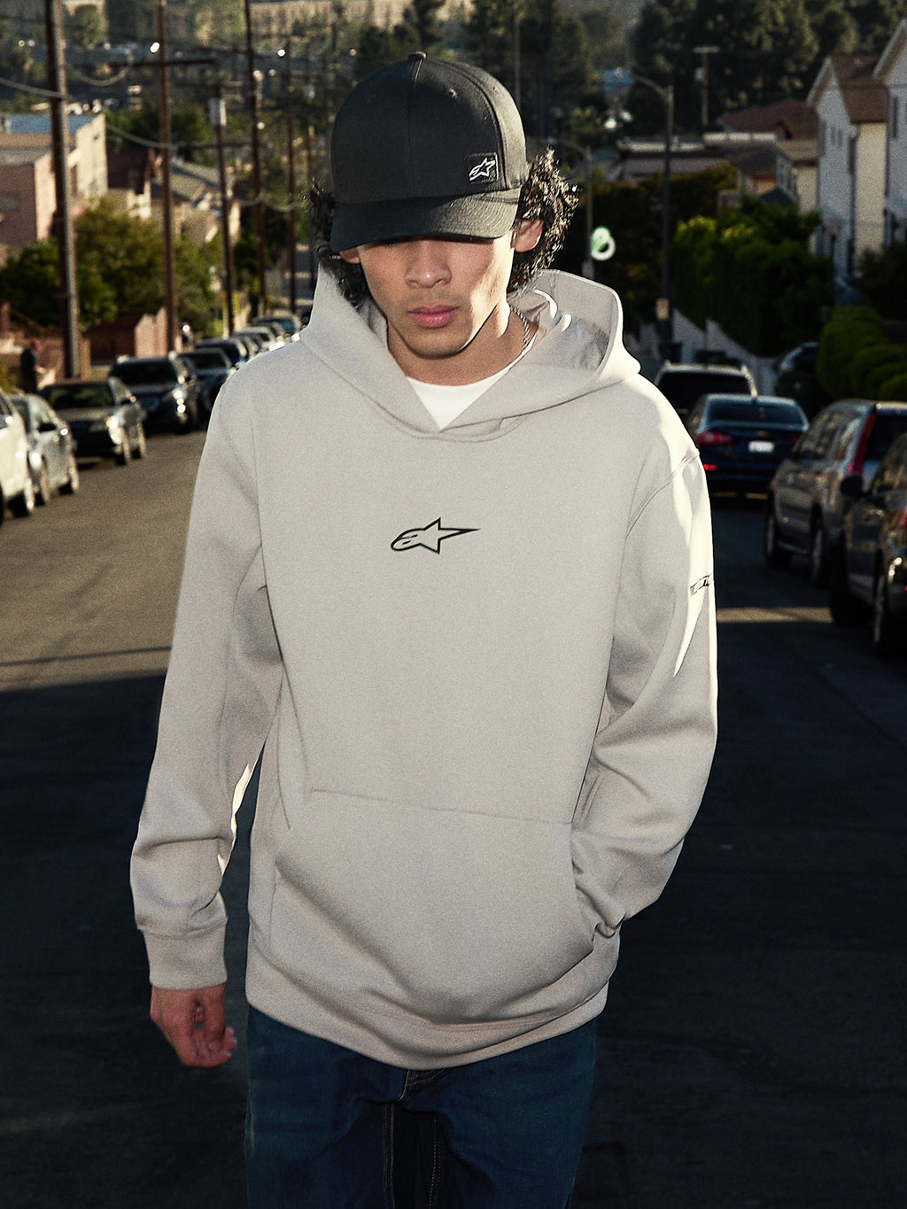 Alpinestars Insight Hoodie, casual lifestyle streetwear, cementgrijs met zwart Astars-logo op de borst, pullover-stijl met kangoeroezak, gedragen met een zwarte pet in een woonstraat, modern motorsport-geïnspireerd ontwerp