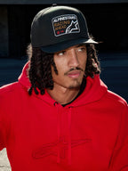 Man met Alpinestars Intuitive Snapback pet in het zwart, voorzien van een gestructureerde kroon en 'Racing Ahead' logo-borduurpatch, gecombineerd met een felrode hoodie, casual lifestyle streetwear