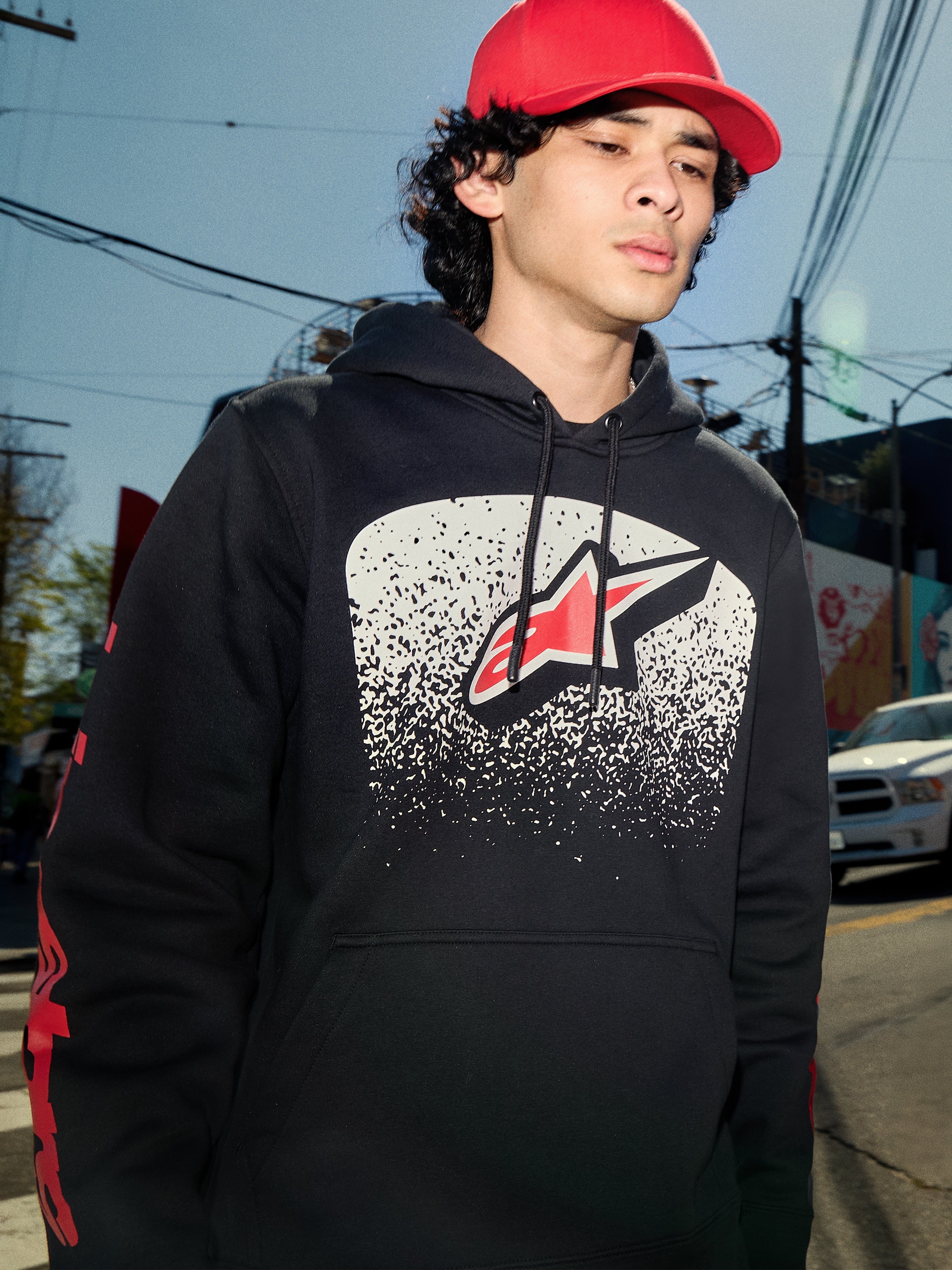 Sweat à capuche Alpinestars Obsessed, polaire noire à enfiler, graphisme blanc avec dégradé MX sur la poitrine et logo étoile rouge, logos rouges sur les manches, streetwear décontracté d'inspiration sport automobile porté dans un environnement urbain.