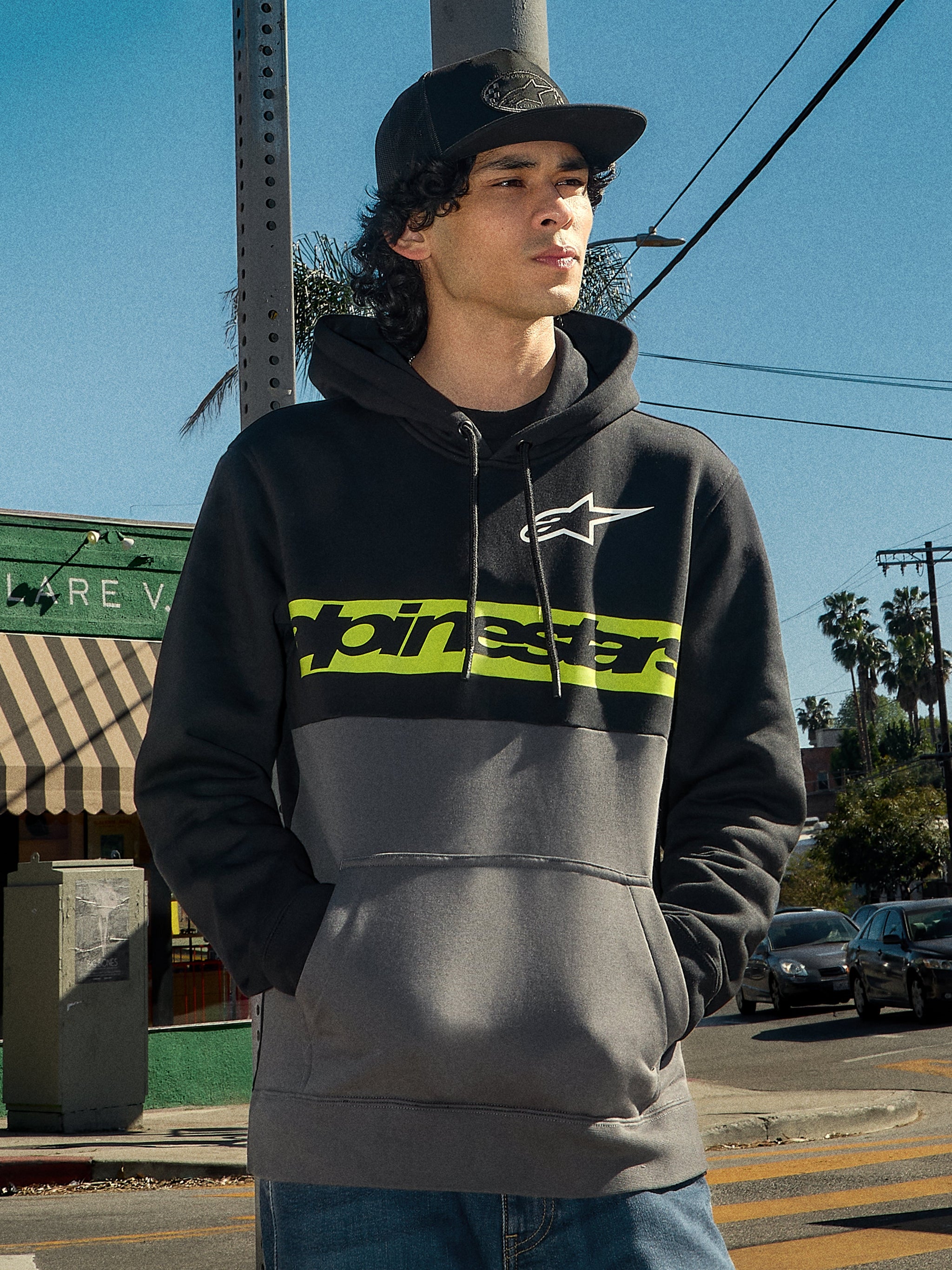 Alpinestars Parallel Hoodie, lifestyle streetwear, color-blocked ontwerp in zwart en gunmetal, limoengroene streep op de borst met Alpinestars-logo, wit Astars-logo, capuchon met trekkoord, gedragen door een man in een stedelijke buitenomgeving.