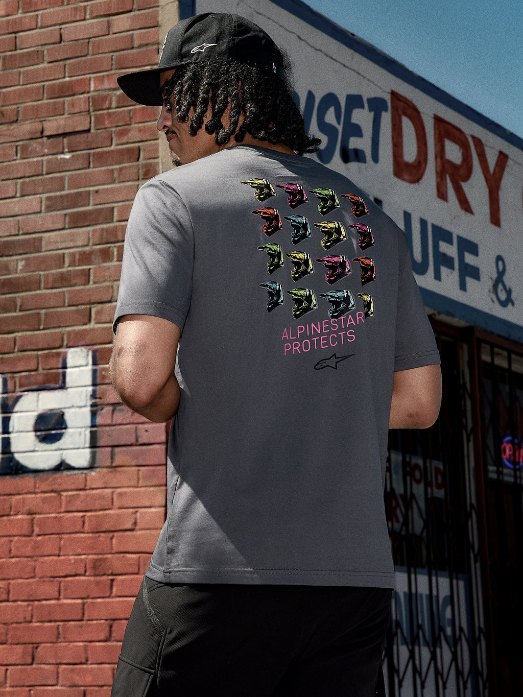 T-shirt Alpinestars Pop Helmet CSF, t-shirt à manches courtes, gris anthracite, graphisme au dos représentant des casques M10 colorés et le texte rose « ALPINESTAR PROTECTS », streetwear décontracté inspiré du sport automobile