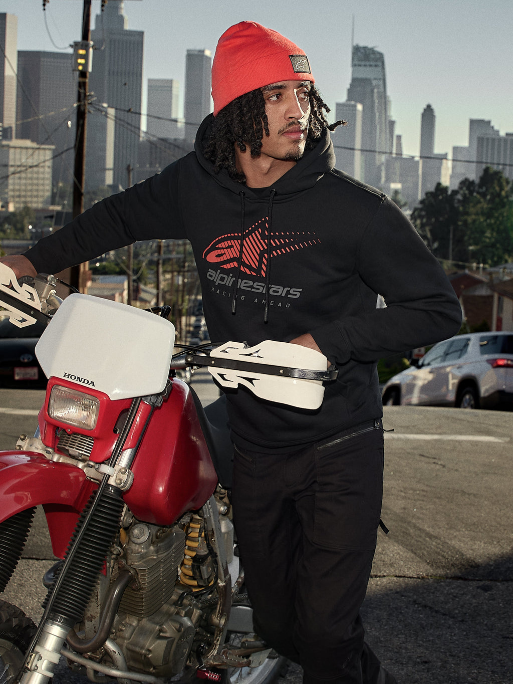Alpinestars Pulse Hoodie, zwarte fleece pullover met capuchon met rood en grijs Astars Combo Logo, gedragen door een man met een oranje muts naast een rode Honda motorfiets in een stedelijke omgeving, casual motorsport streetwear