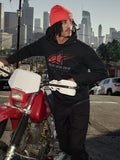 Alpinestars Pulse Hoodie, zwarte fleece pullover met capuchon met rood en grijs Astars Combo Logo, gedragen door een man met een oranje muts naast een rode Honda motorfiets in een stedelijke omgeving, casual motorsport streetwear